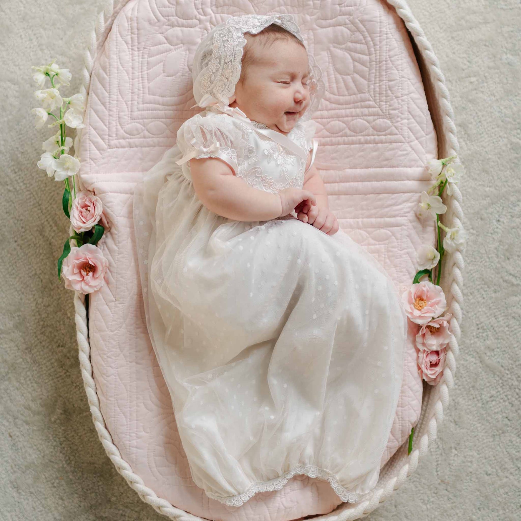 A baby sleeps in a bassinet adorned with flowers, dressed in the Sophia Layette with a lace bonnet and flutter sleeves.