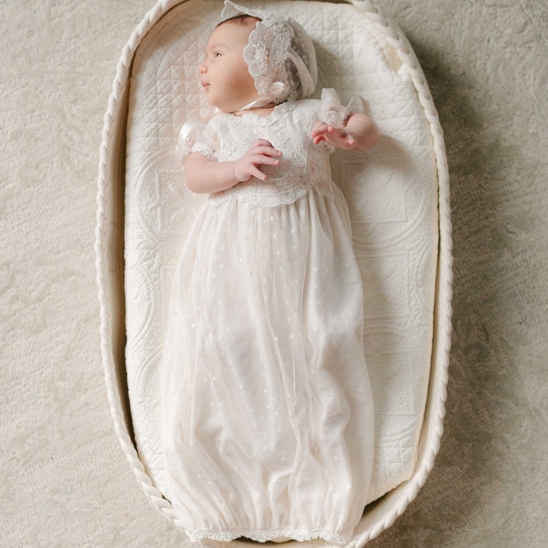 A newborn lays in a woven bassinet, dressed in the Sophia Layette with a matching lace bonnet, surrounded by soft textures.