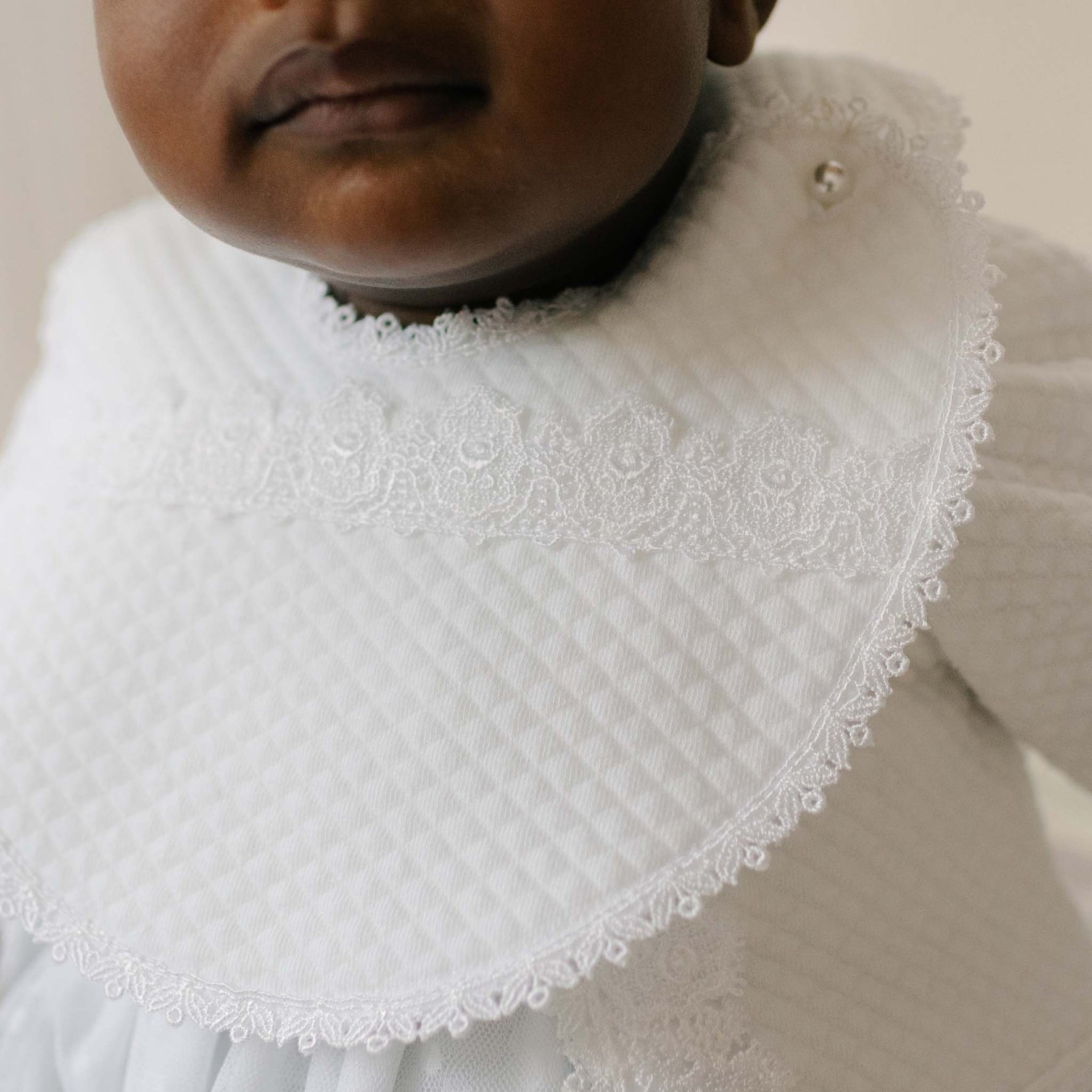 Close-up of a baby wearing the Stella Bib. Details of the bib include plush quilted cotton with a white edge lace trim and pearl style button enclosure. Perfect for unique baby gifts.
