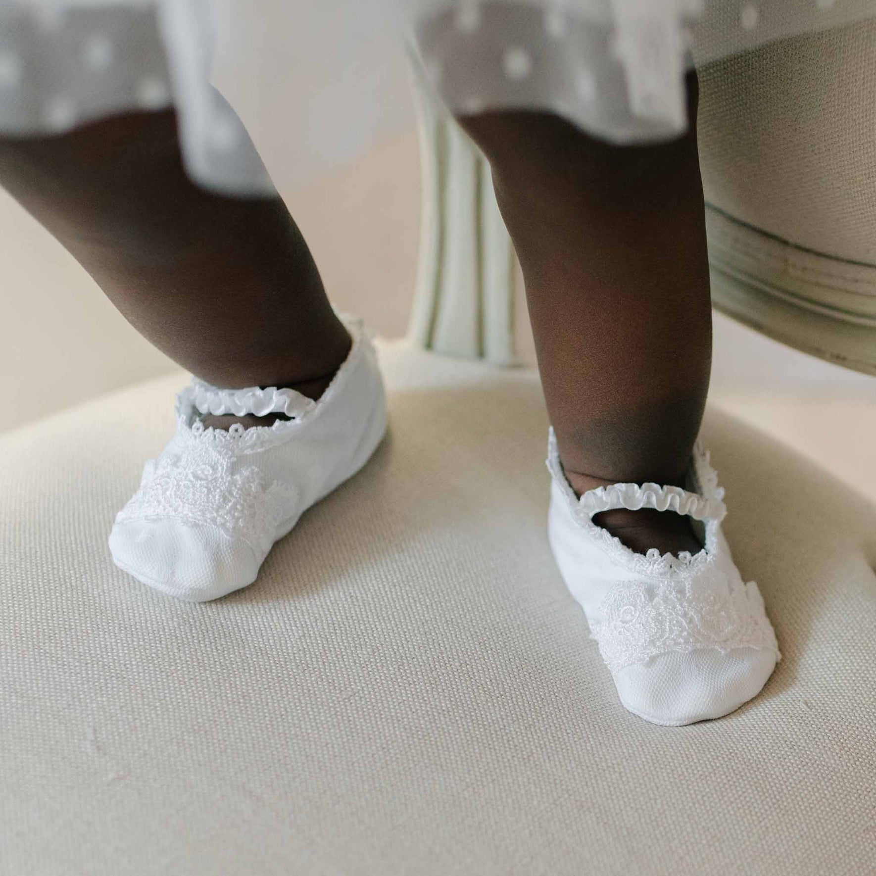 Close up of a baby wearing a pair of the Stella Booties and standing on the seat of a beige chair. Features of the booties include white pima cotton with a netting overlay and white Venice lace. 
