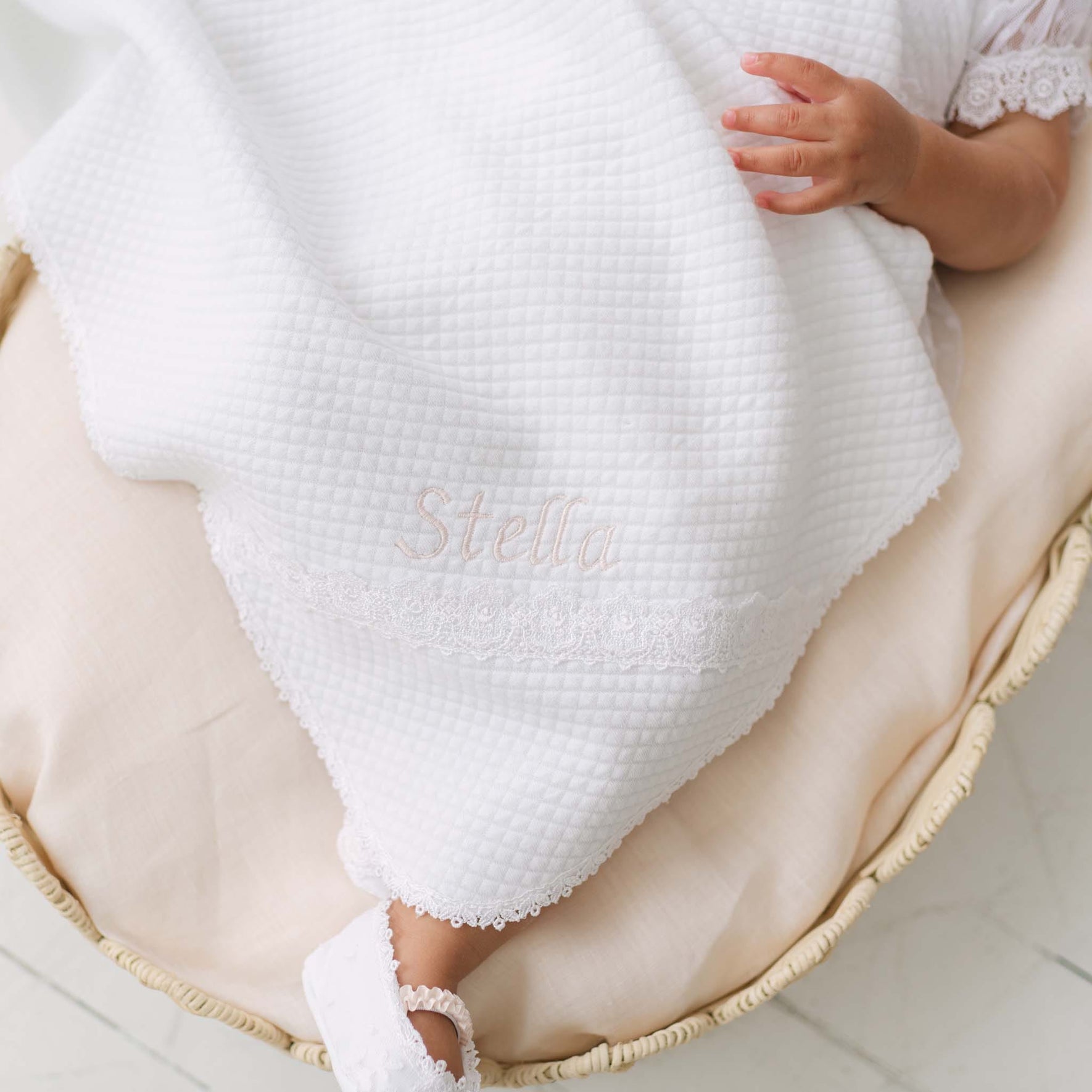 Close up photo of the Stella Personalized Blanket covering a baby in a wicker bassinet. Details show the corner of the blanket embroidered with the name "Stella" and trimmed with white edge lace. Part of Christening Gowns' high end baby blankets.
