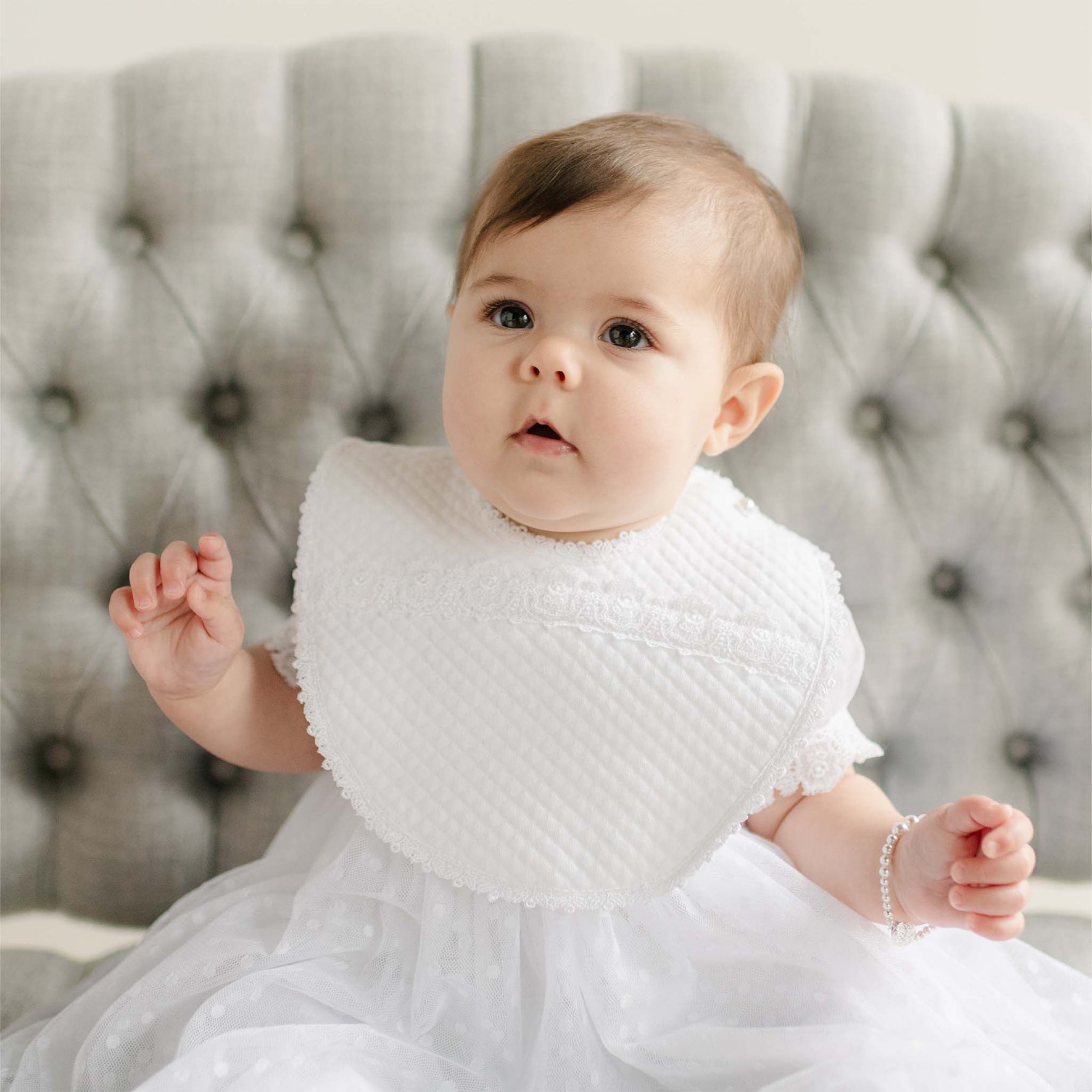 Baby wearing the Stella Bib against a gray tufted background. Details of the bib show a quilted cotton with a white edge lace trim. Perfect for unique baby gifts.