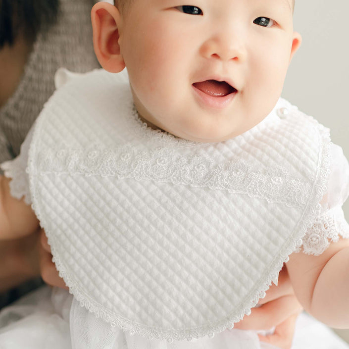 Baby wearing the Stella Bib, held by an adult in the background. The bib features quilted cotton with white Venice lace.