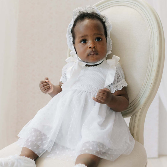 Baby in a white dress and bonnet sitting on a white chair.