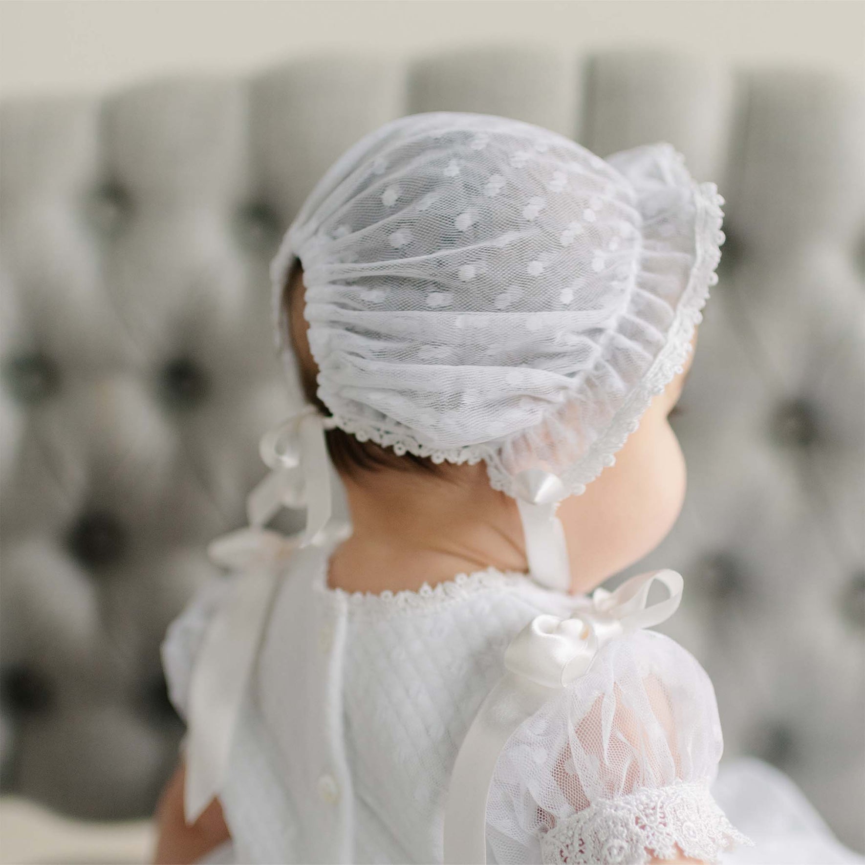 Back details of the Stella Lace Bonnet as worn by a baby against a blurred background.