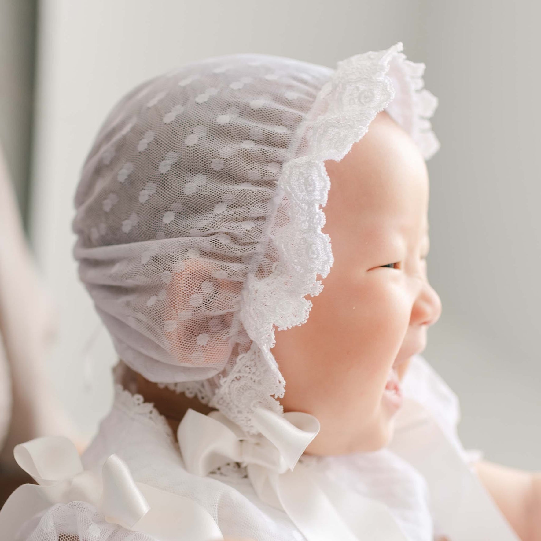 Baby wearing the Stella Lace Bonnet against a soft focus background. The bib features a white dotted lace and a white edge lace trim with silk ribbon ties. Part of Christening Gowns' high end baby clothes.