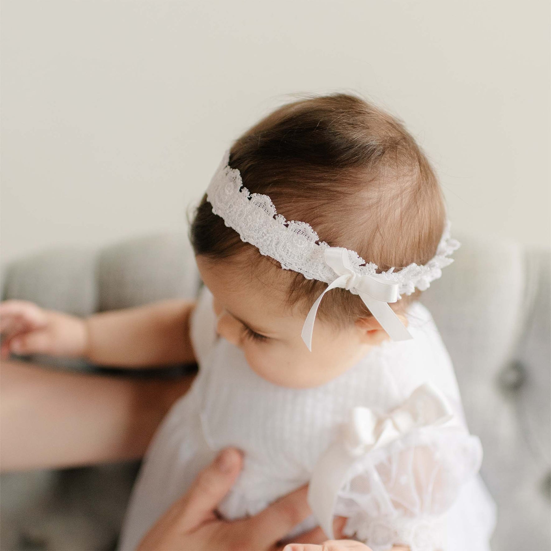 Close up of a baby wearing the Stella Lace Headband and matching Dress against a blurred background. Details show the white Venice lace and silk ribbon bow. 