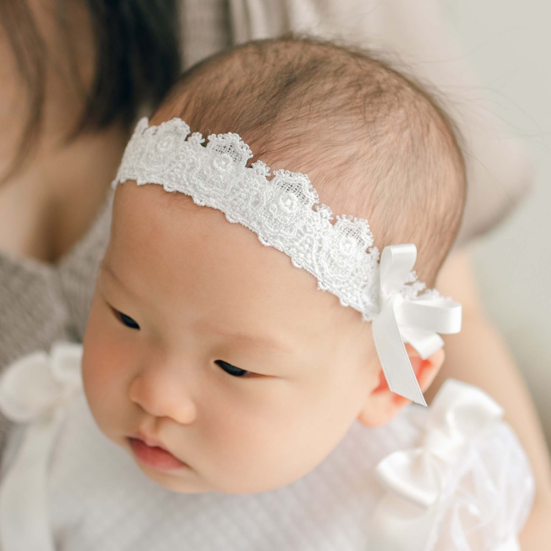 Baby wearing the Stella Lace Headband and looking to the side as a woman in the background appears to hold her. Details of the headband include a Venice lace band with elastic and a silk ribbon bow.