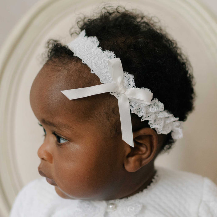Baby wearing the Stella Lace Headband, looking to the side. Details of the headband include a Venice lace band with elastic and a silk ribbon bow. Part of Christening Gowns' high end baby clothes.