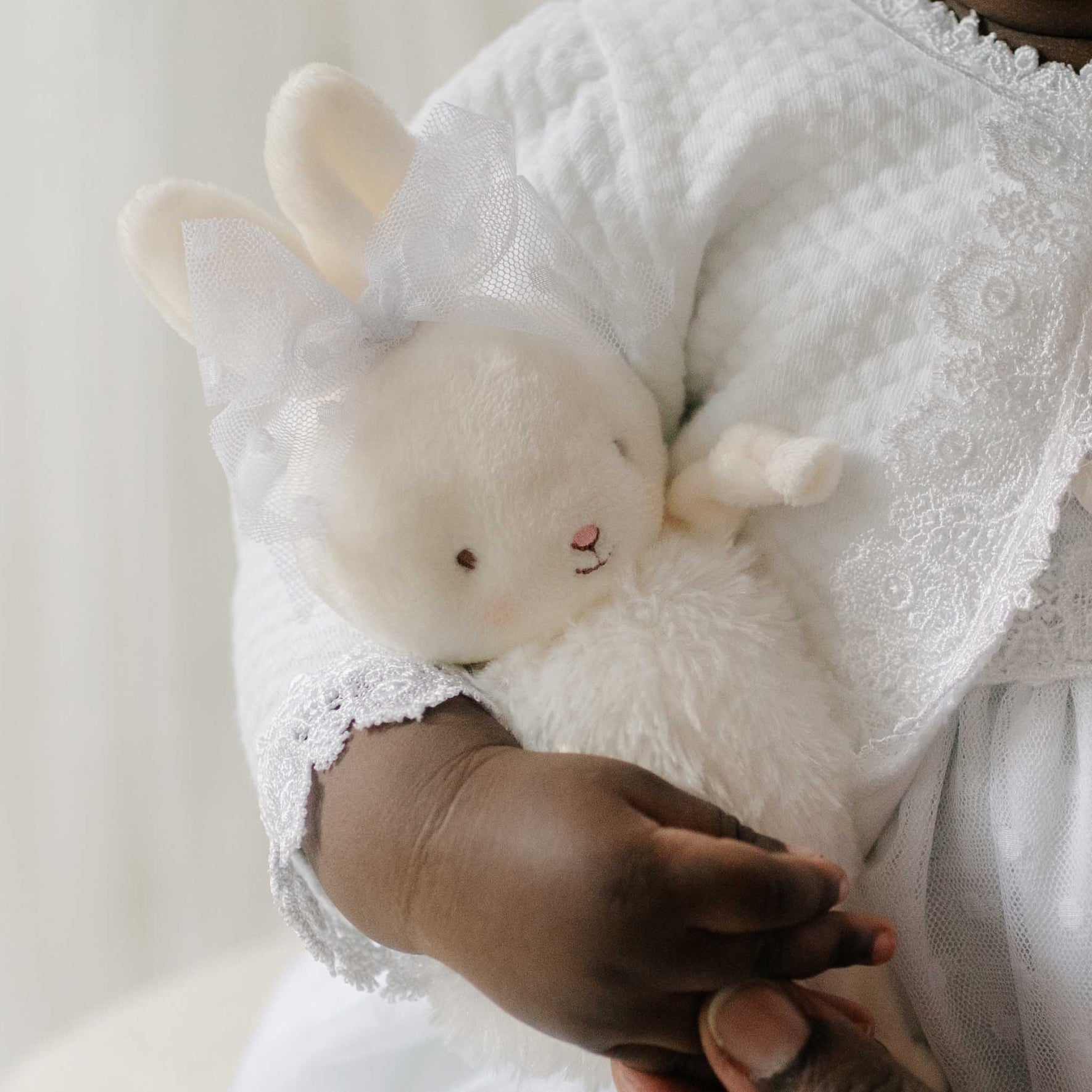 Close up of a baby holding the Stella Roly Poly Bunny against a soft white background. The plush bunny includes a Stella lace bow tied around its ears. Perfect for a baptism gift from godparents.