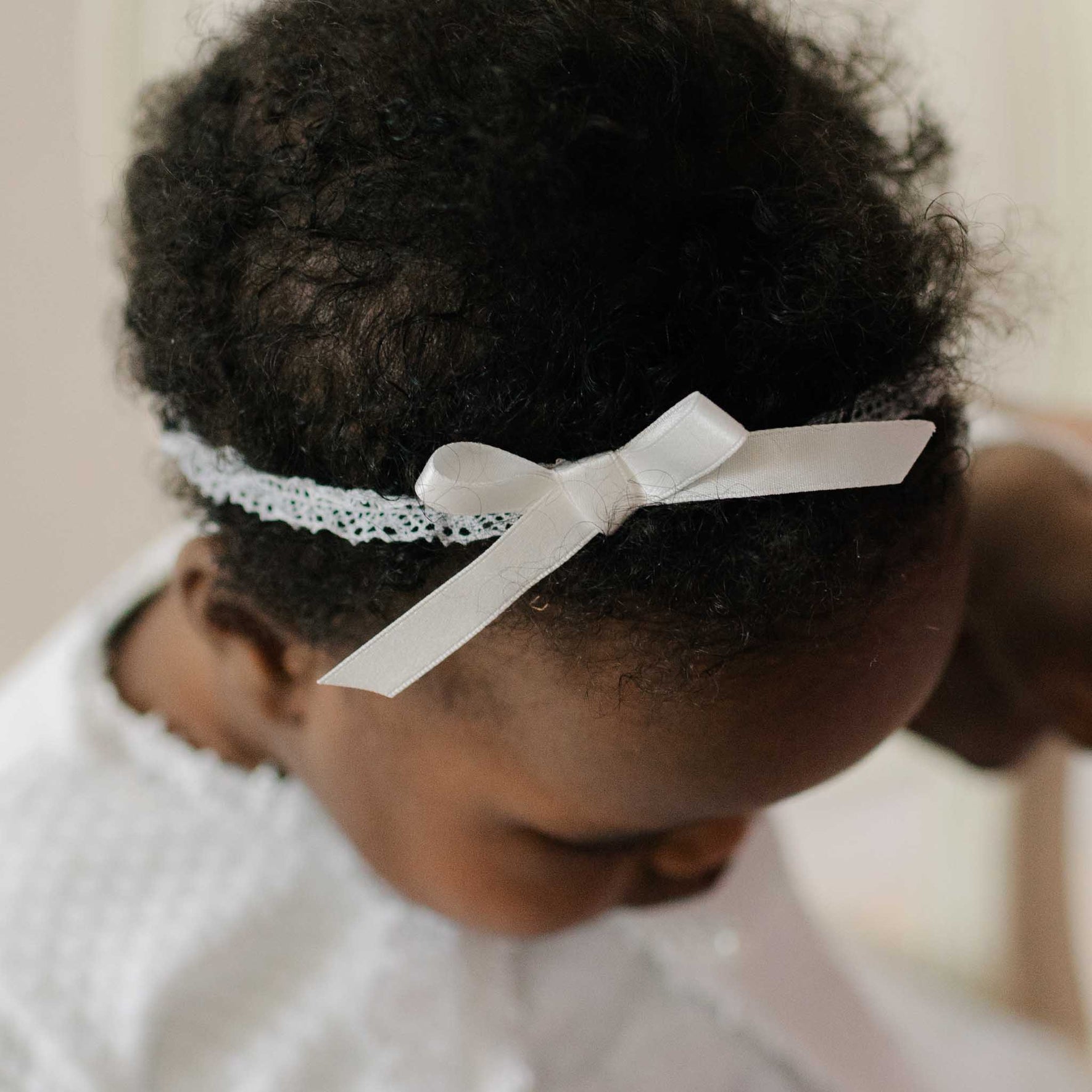 Close up photo of a baby wearing the Stella Lace Headband, looking down. Details show soft white Venice lace with a silk ribbon bow. Part of Baby Beau & Belle's luxury baby clothes.