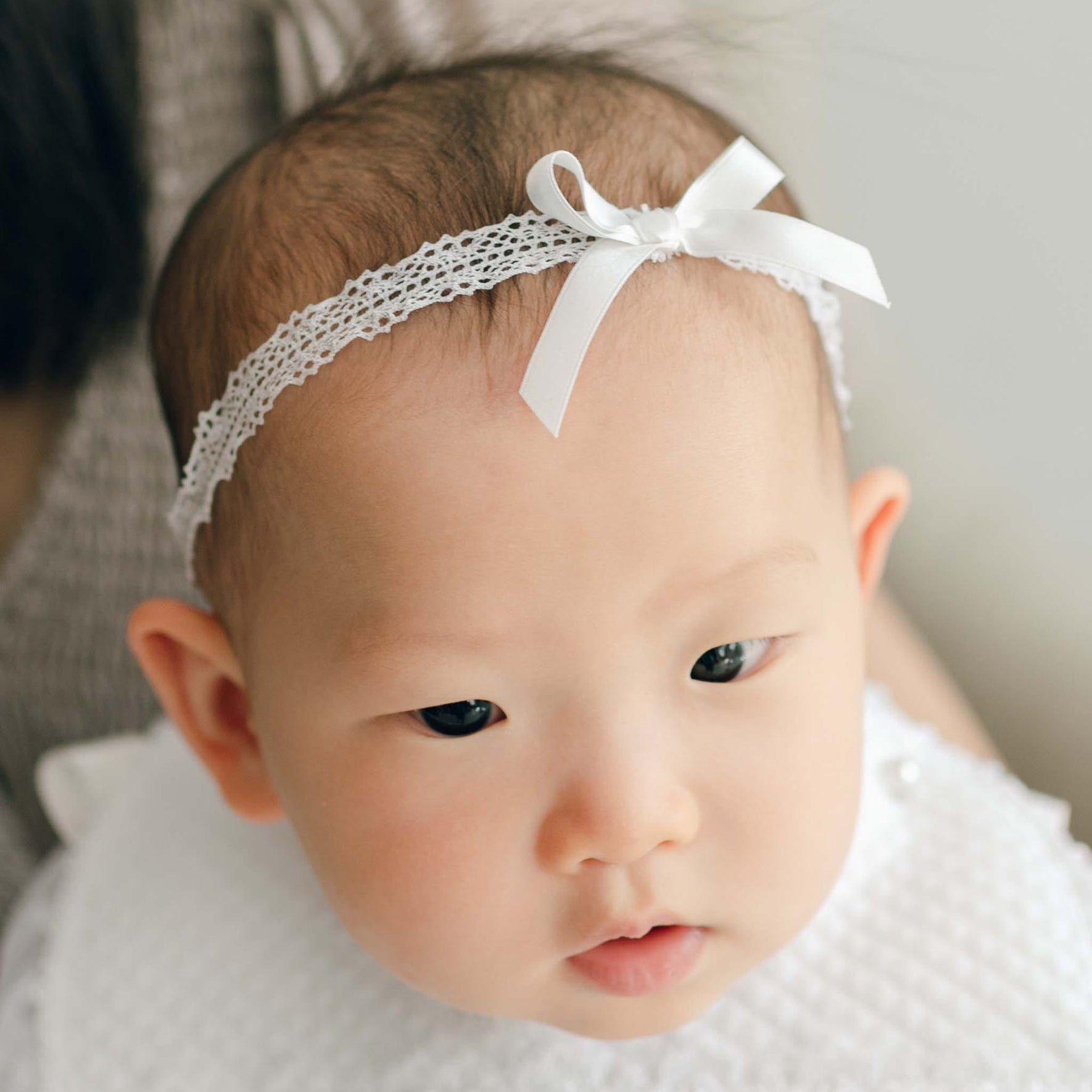 Newborn baby wearing the Stella Silk Ribbon Headband and matching Bib. The headband features white Venice lace and a silk ribbon bow. A woman appears to be holding her outside of the frame.