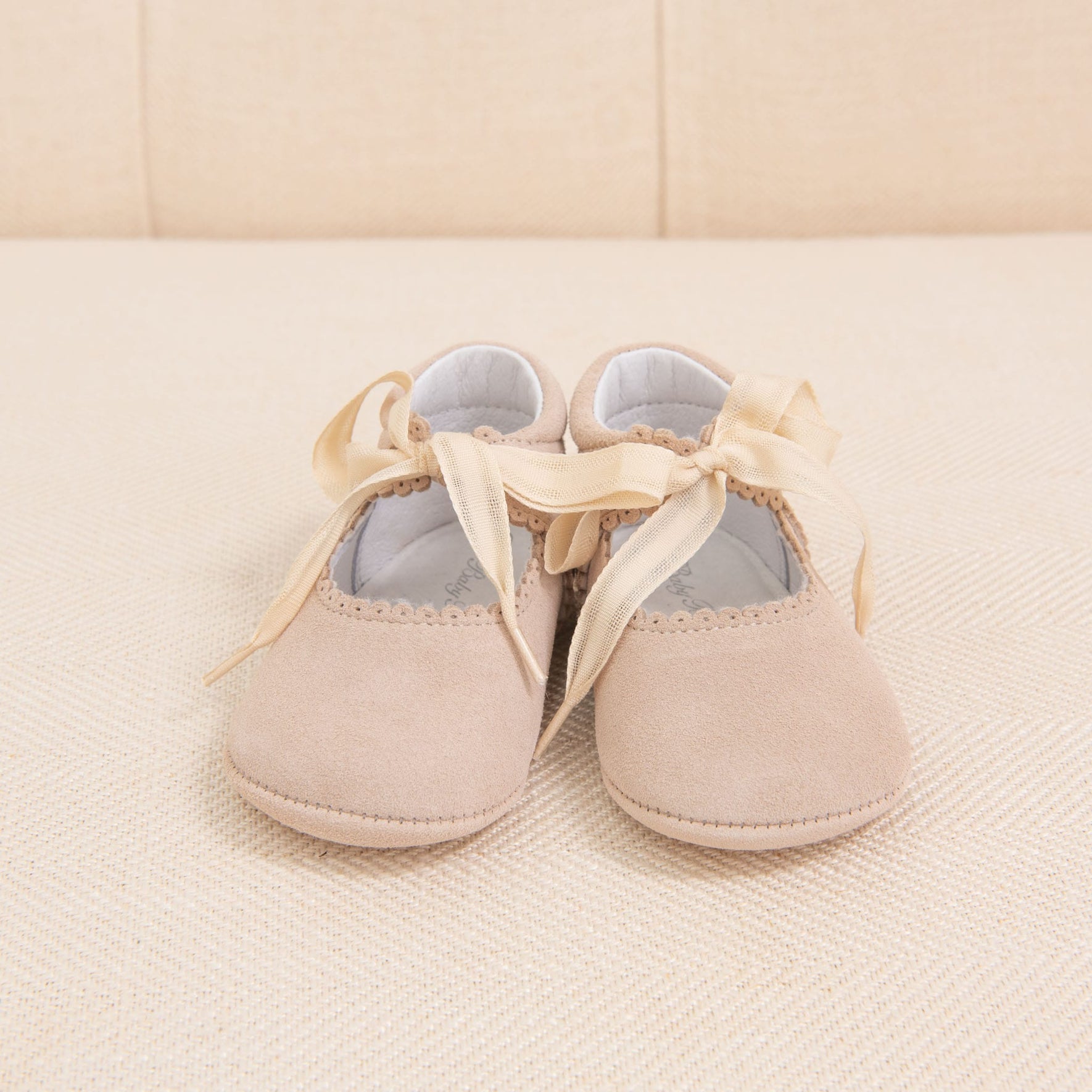 A pair of Tan Suede Tie Mary Janes by Baby Beau & Belle, crafted from soft suede leather and adorned with satin ribbons tied into bows, is placed on a cream-colored textured surface. The handmade shoes feature subtle scalloped edges along the opening.