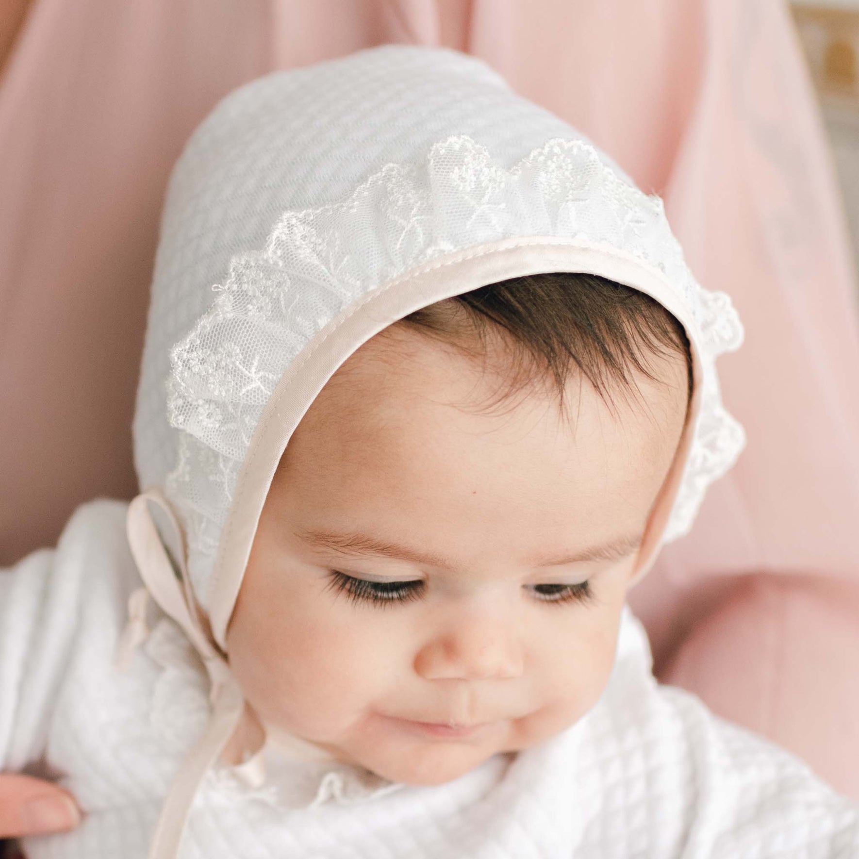 Newborn baby girl wearing the Tessa Quilt Christening Bonnet made from a quilted cotton in white and trimmed in pink champagne silk and light ivory floral lace.