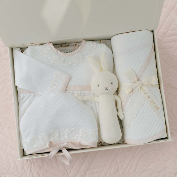 A neatly arranged Tessa Newborn Gift Set - Save 10% contains baby clothes including a pima cotton newborn coming home gown, a plush bunny toy, and a swaddle blanket. The clothing and blanket are primarily white with light pink accents and delicate Venice lace. The box has a quilted pattern background that matches the items inside.