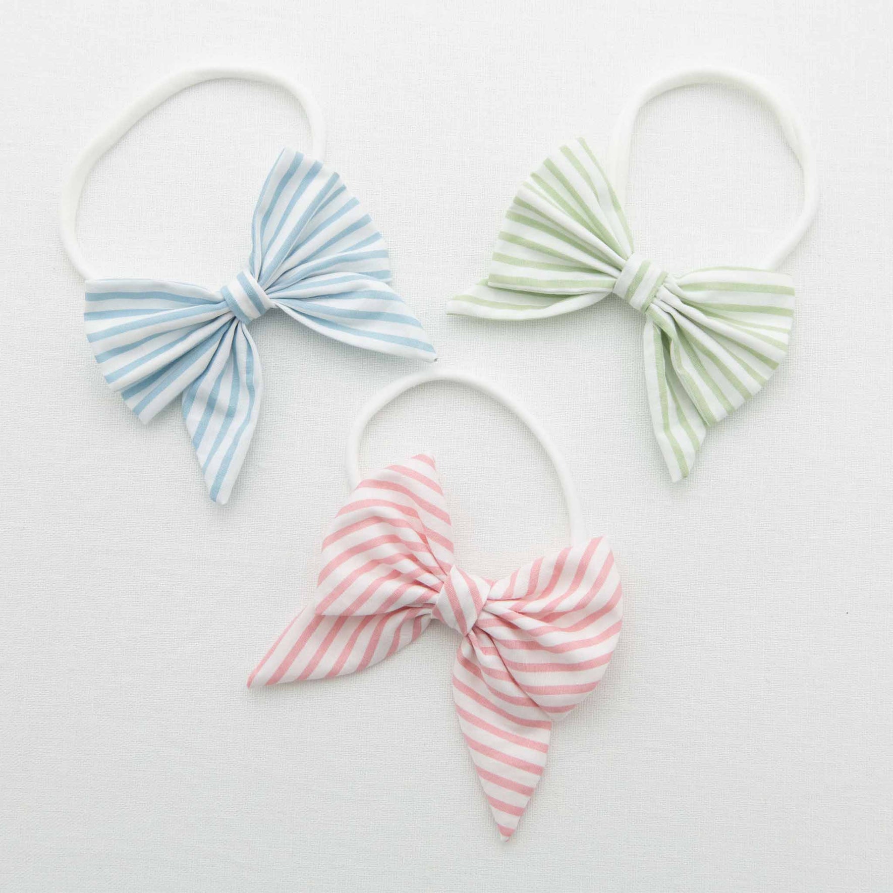 Three Thea Bow Headbands with handmade bows are arranged on a white surface. One is blue and white, another is green and white, and the third is pink and white.