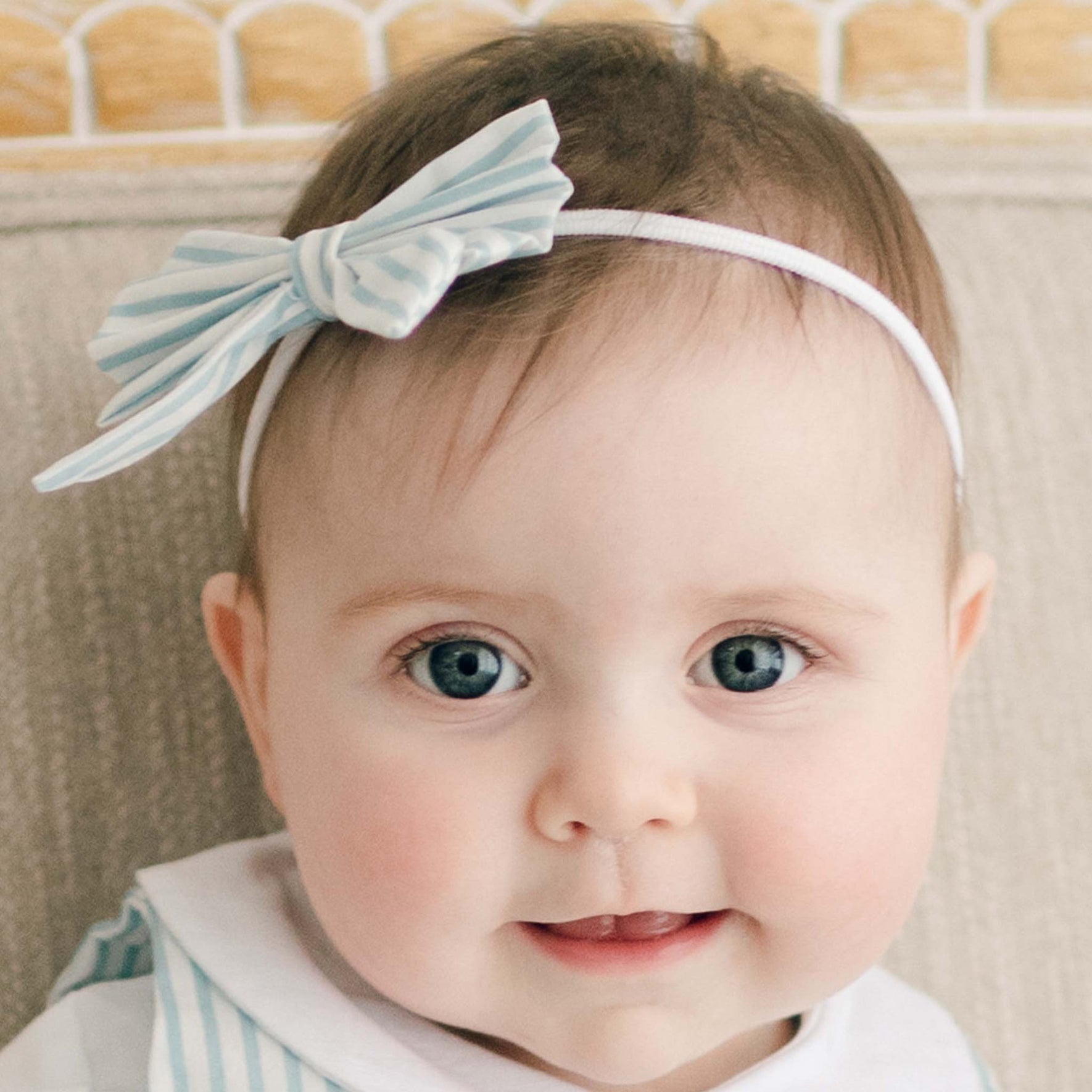 A baby wearing Thea Bow Headband, which features a white and blue striped design crafted from hand-stretched nylon, and a matching striped outfit is sitting against a neutral-colored background, looking forward.
