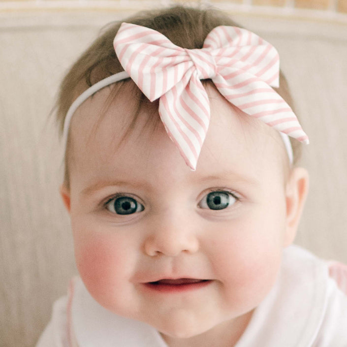A baby with big eyes and rosy cheeks is looking at the camera, wearing a white outfit complemented by Thea Bow Headband, which features a handmade bow in pink and white stripes. The background is softly blurred.