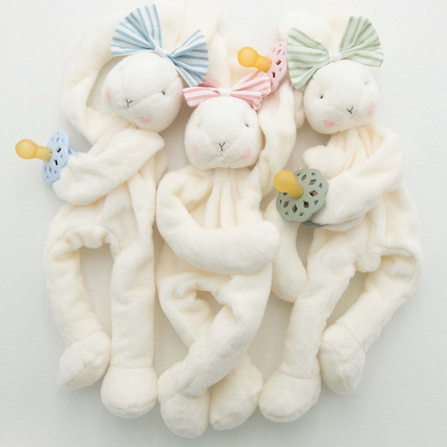Three Thea Bunny Buddy pacifier holders with soft white fur are positioned side by side, each clutching a colorful pacifier in blue, pink, and green. Decorated with pastel-striped bows on their heads, they feature a cozy and relaxed posture—ideal as a baby-safe christening gift.