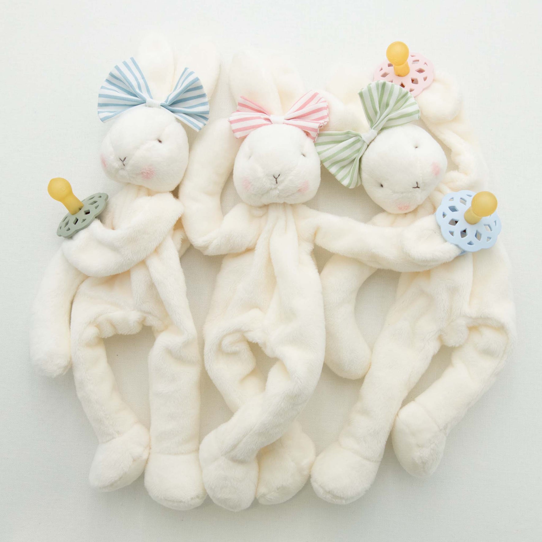 Three Thea Bunny Buddy pacifier holders lie side by side, ideal as a baby-safe christening gift. Each features a cream-colored body with a pastel bow on its head and holds a pacifier in green, blue, or pink. Their eyes and noses are delicately embroidered.
