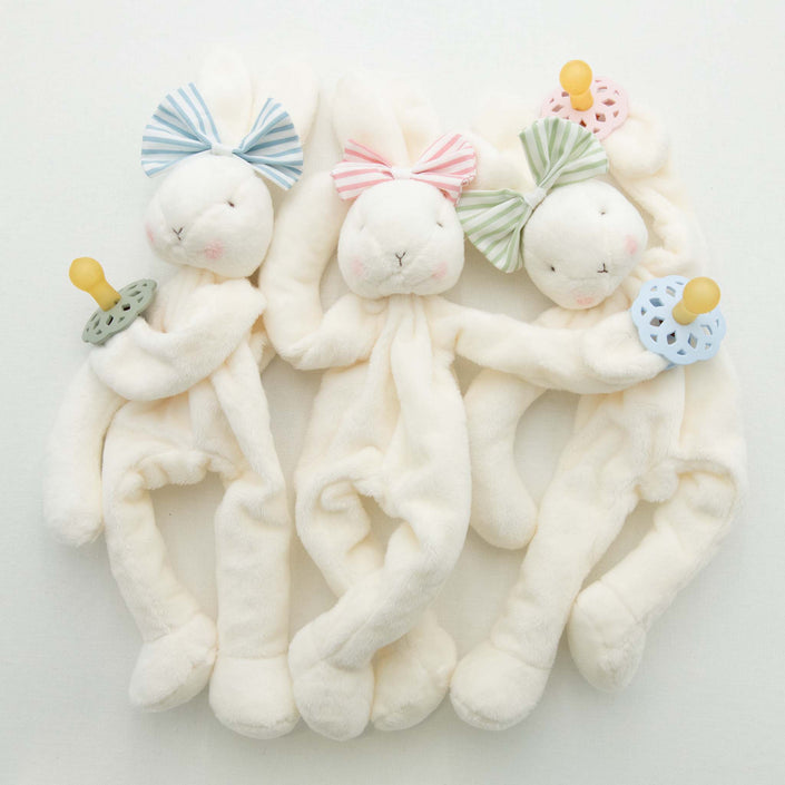 Three Thea Bunny Buddy pacifier holders lie side by side, ideal as a baby-safe christening gift. Each features a cream-colored body with a pastel bow on its head and holds a pacifier in green, blue, or pink. Their eyes and noses are delicately embroidered.