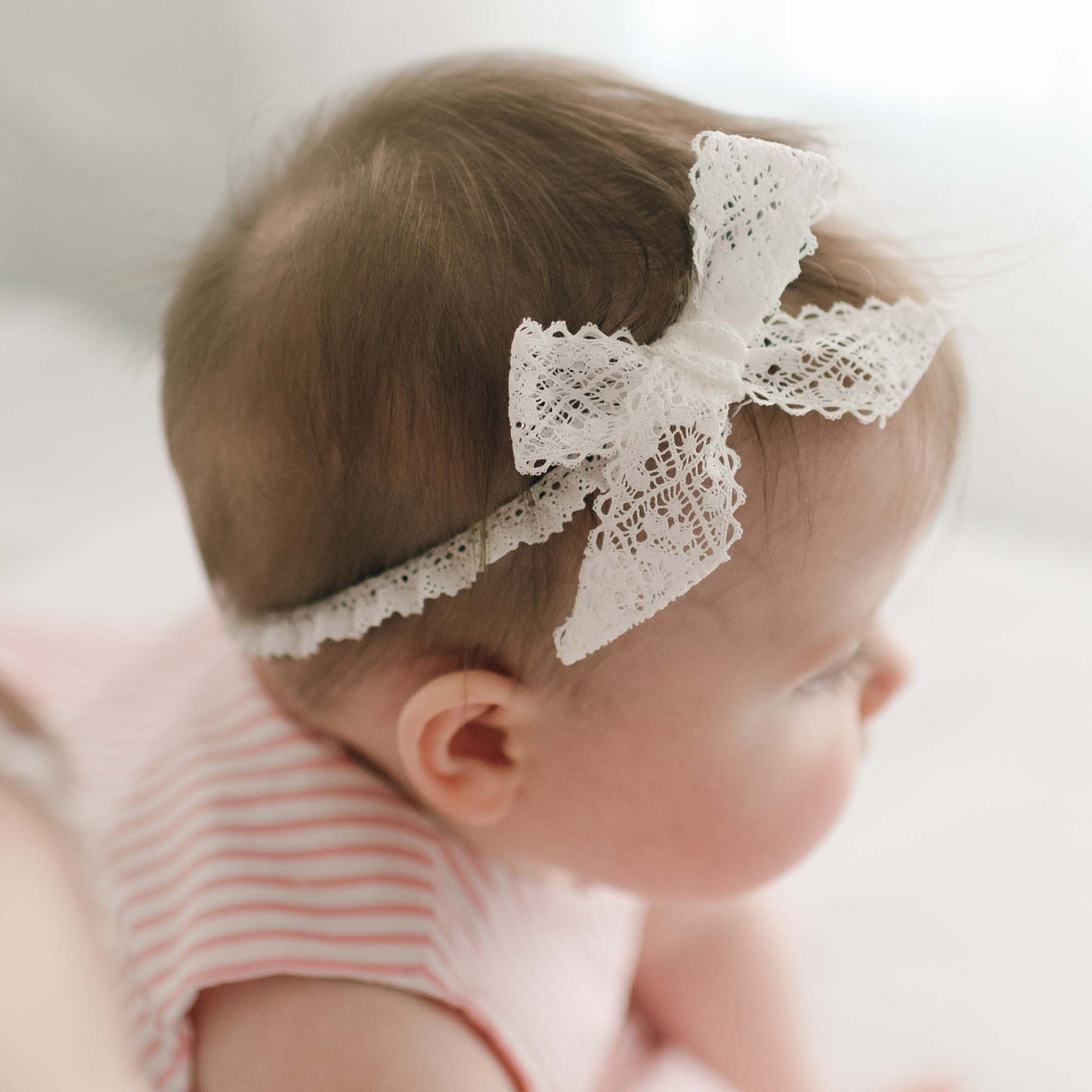 A baby wearing Thea Lace Headband, handmade in the USA, lies on a soft surface. Dressed in a pink and white striped outfit, the baby gazes to the side against a softly blurred background.