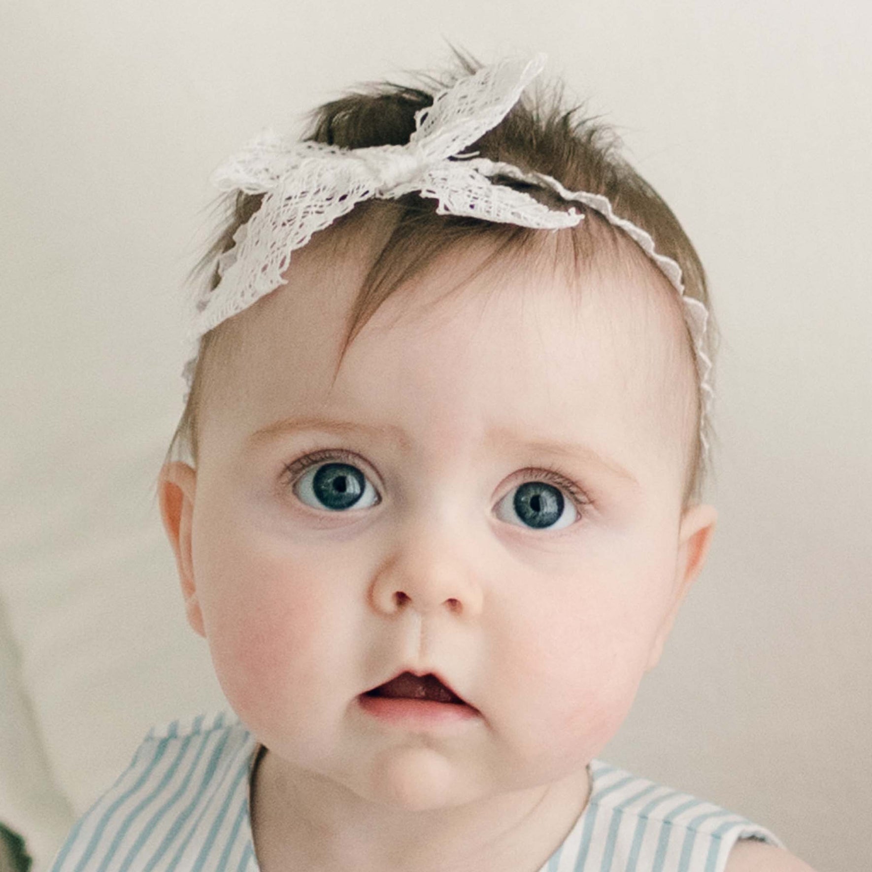 The baby with blue eyes and light hair is wearing Thea Lace Headband, a white stretch lace headband featuring a bow, handmade in the USA. Dressed in a sleeveless, light blue and white striped outfit, the baby gazes directly at the camera against a soft, neutral background.