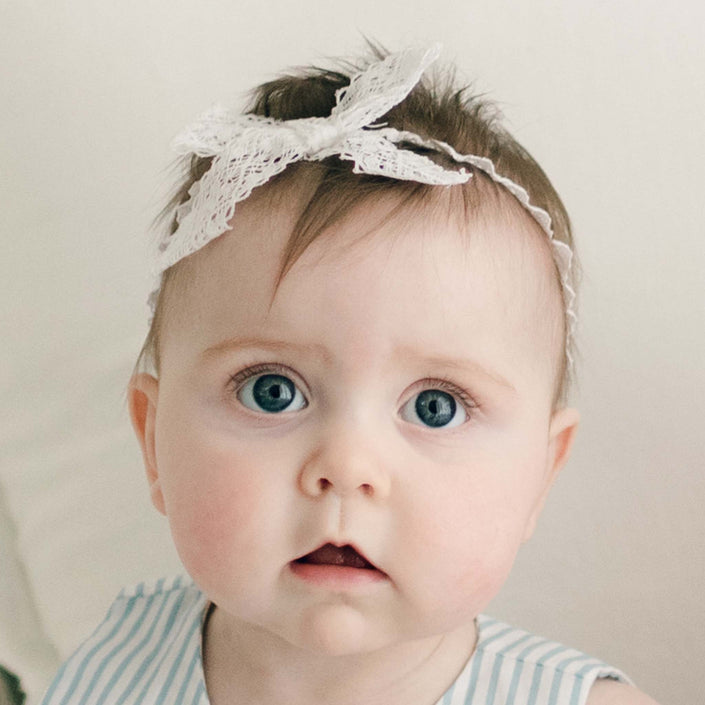 The baby with blue eyes and light hair is wearing Thea Lace Headband, a white stretch lace headband featuring a bow, handmade in the USA. Dressed in a sleeveless, light blue and white striped outfit, the baby gazes directly at the camera against a soft, neutral background.