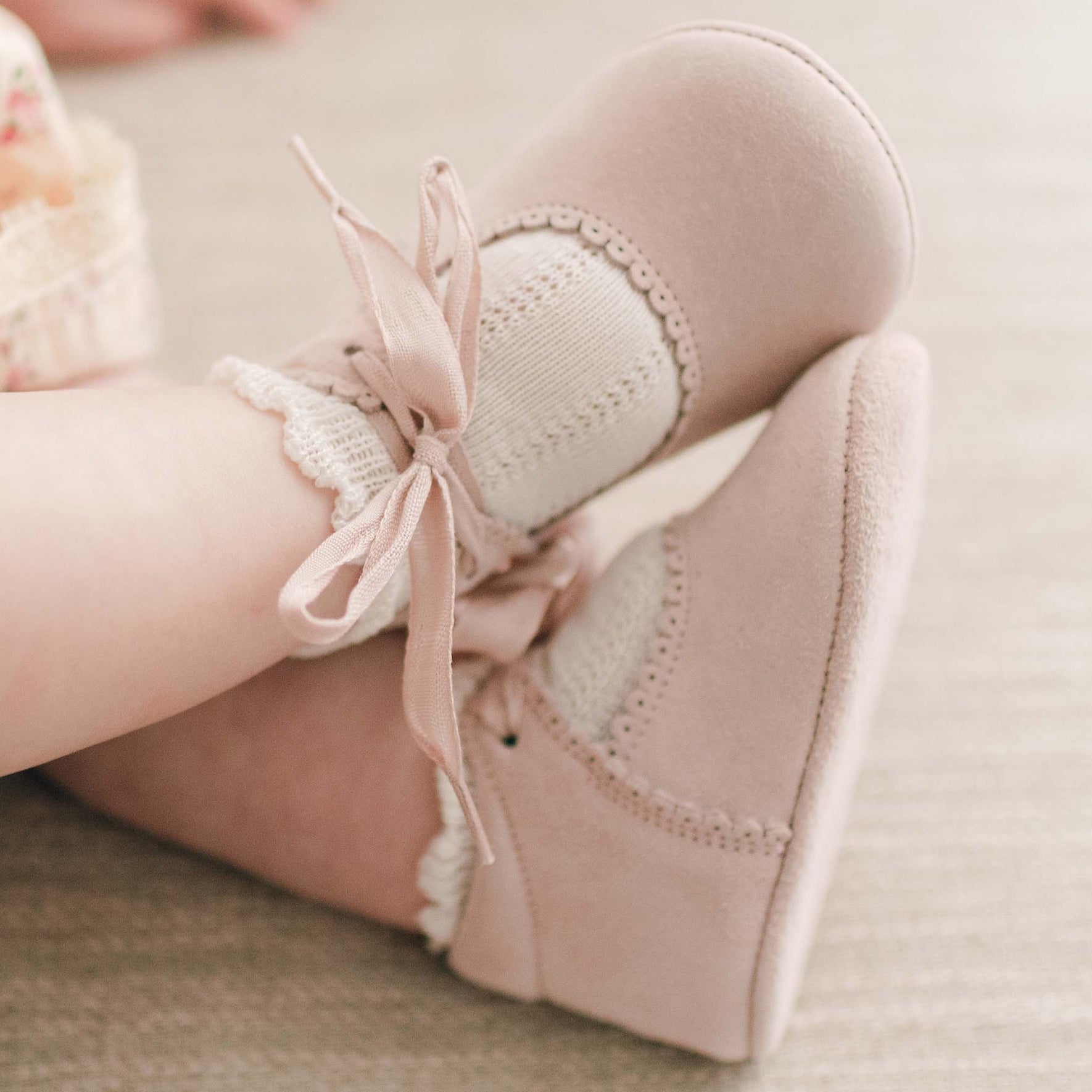 A close-up image of an infant's feet wearing soft pink Thea Suede Tie Mary Janes from Baby Beau & Belle. The handmade shoes feature matching pink ribbon ties and are paired with white knitted socks that have a delicate lacy frill on the cuffs.
