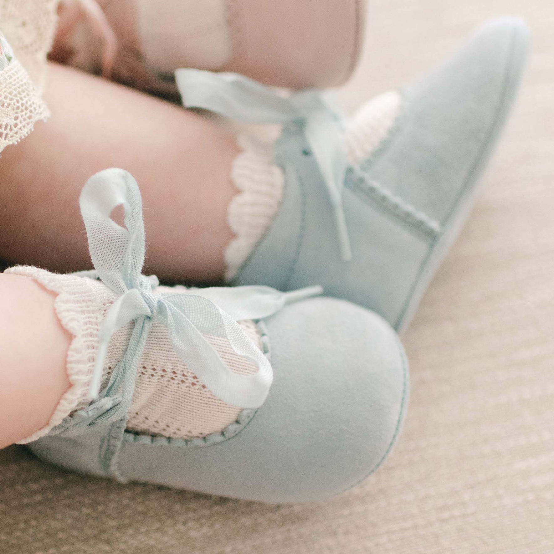 Close-up of a baby's feet wearing light blue, soft-soled, handmade Thea Suede Tie Mary Janes with ribbon laces. The baby is also wearing white, lace-trimmed socks. These adorable shoes by Baby Beau & Belle rest on a neutral-colored surface.