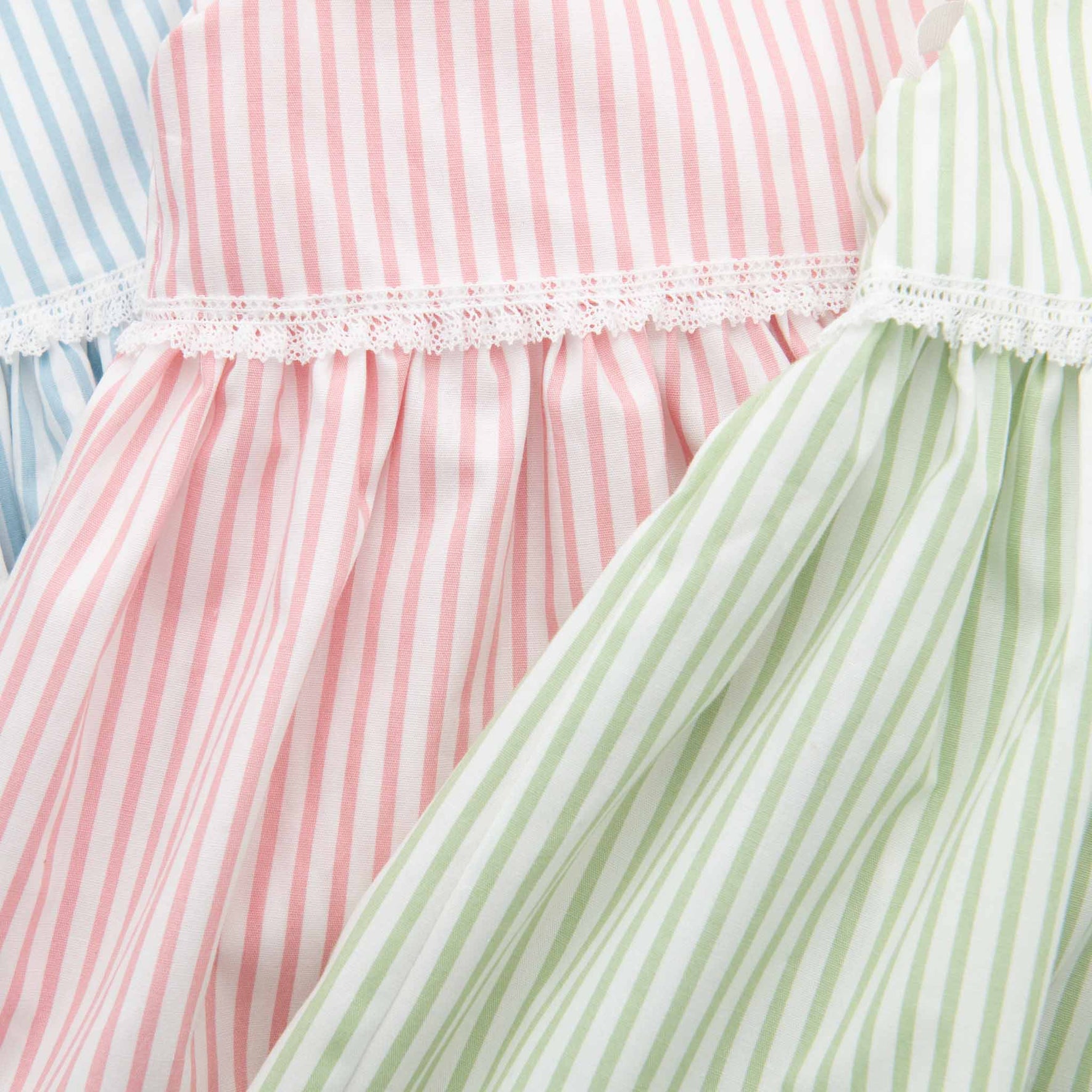 Close-up of three pastel-colored striped cotton fabrics arranged diagonally. The textiles, similar to those used in the Thea Wrap Dress, display vertical stripes in blue, pink, and green, each adorned with a lace trim along the top edge.