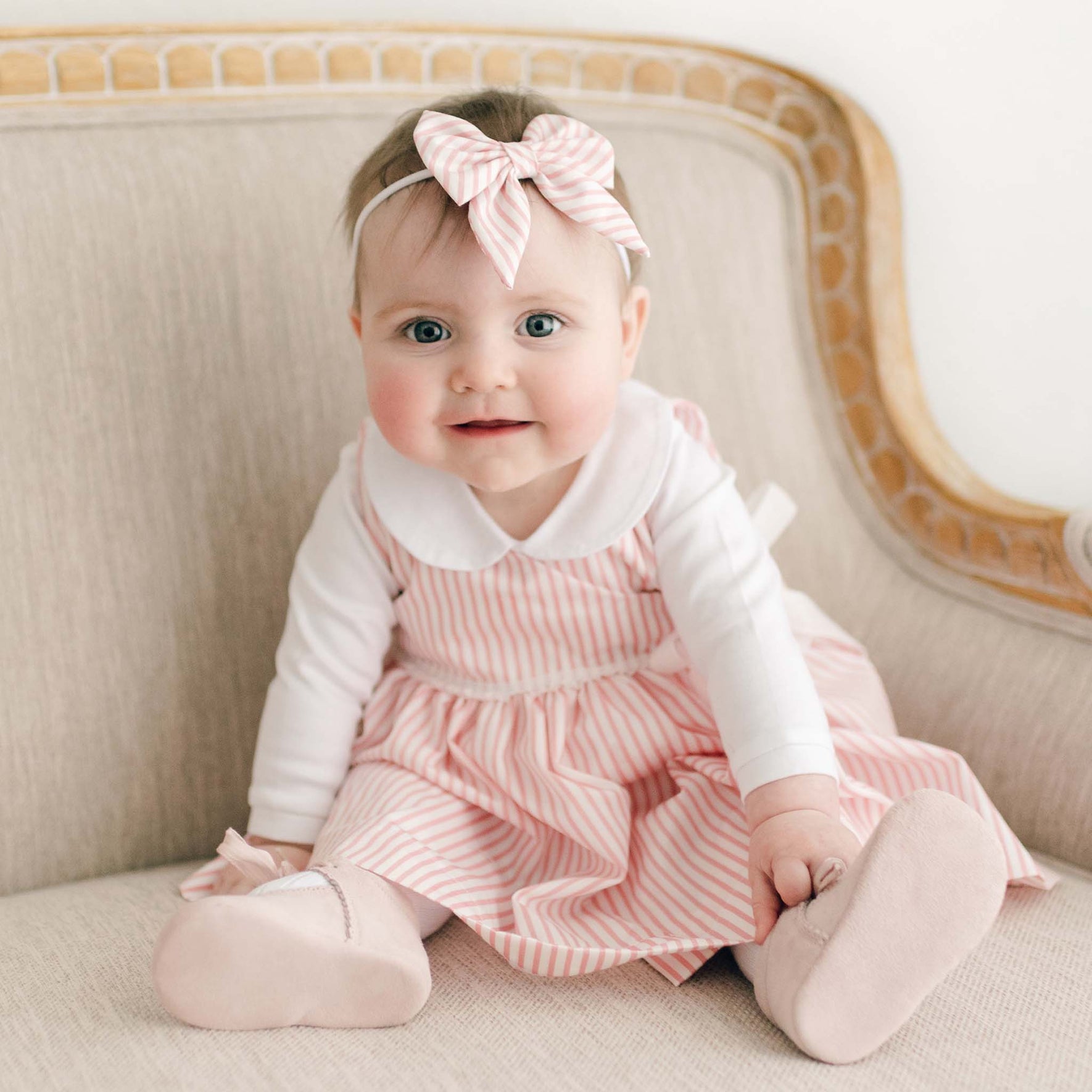 A baby dressed in a Thea Wrap Dress, featuring pink and white stripes and styled with a Thea Bow Headband, sits on a beige cushioned chair. Her look is completed with matching pink shoes, as she gazes forward with her arms gently resting on the chair.