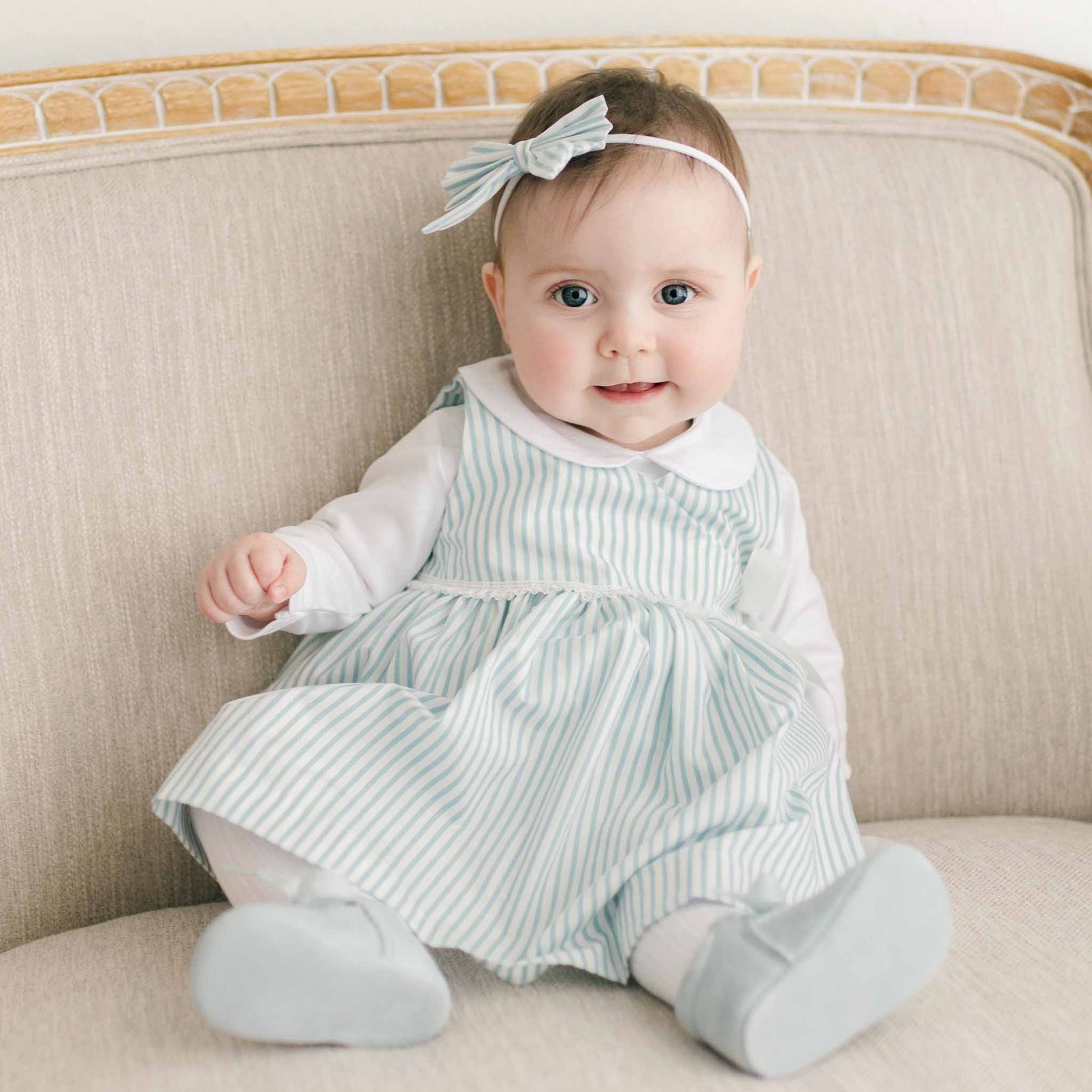 A baby in a light blue and white striped Thea Wrap Dress and a Thea Bow Headband sits on a beige sofa. With hands resting on its lap, the baby gazes forward with a neutral expression. The sofa, framed in light-colored wood, complements the charming ensemble perfectly.