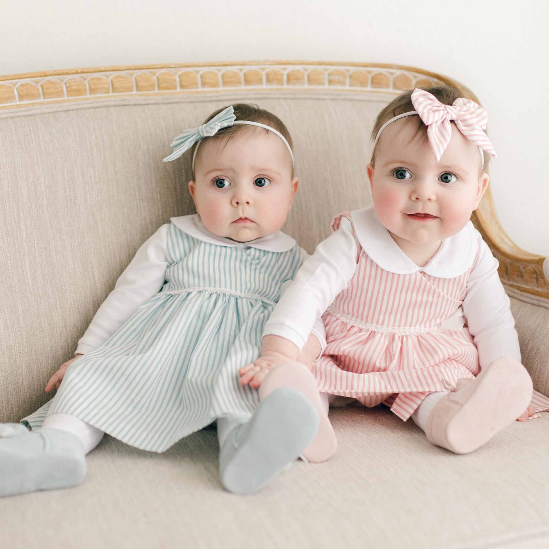 Two babies are sitting on a beige couch, both dressed alike in adorable Thea Wrap Dresses and matching Thea Bow Headbands. One is wearing a blue wrap dress and headband, while the other is clad in pink. They gaze toward the camera with neutral expressions.