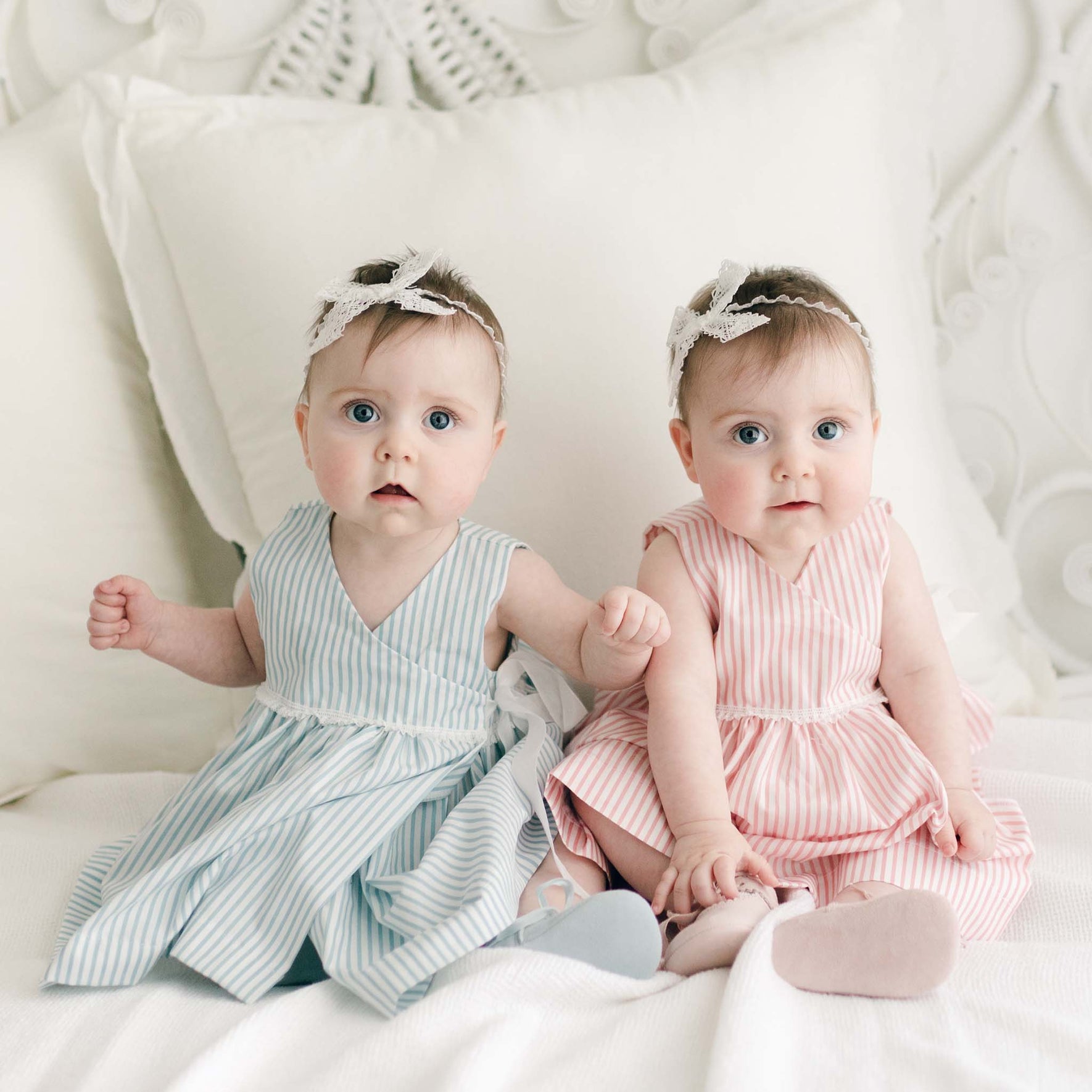Two babies sit on a bed with white pillows, wearing matching striped cotton Thea Wrap Dresses. The baby on the left is dressed in a blue-striped version, while the one on the right sports pink stripes. Both are adorned with charming lace headbands that resemble Thea Bow Headbands, gazing toward the camera with curious eyes.