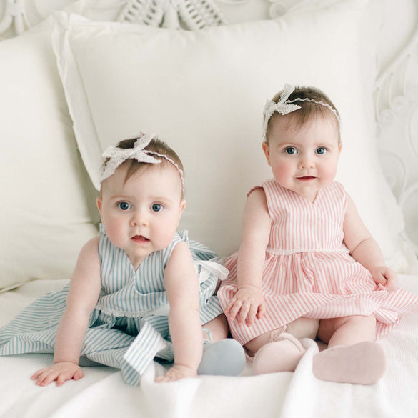 Two babies are sitting on a white bed, dressed in adorable pastel-striped Thea Wrap Dresses. The one on the left is wearing a blue striped wrap dress, while the right dons pink stripes. Both sport matching lace headbands reminiscent of Thea Bow Headbands. Soft white pillows complement the serene scene.