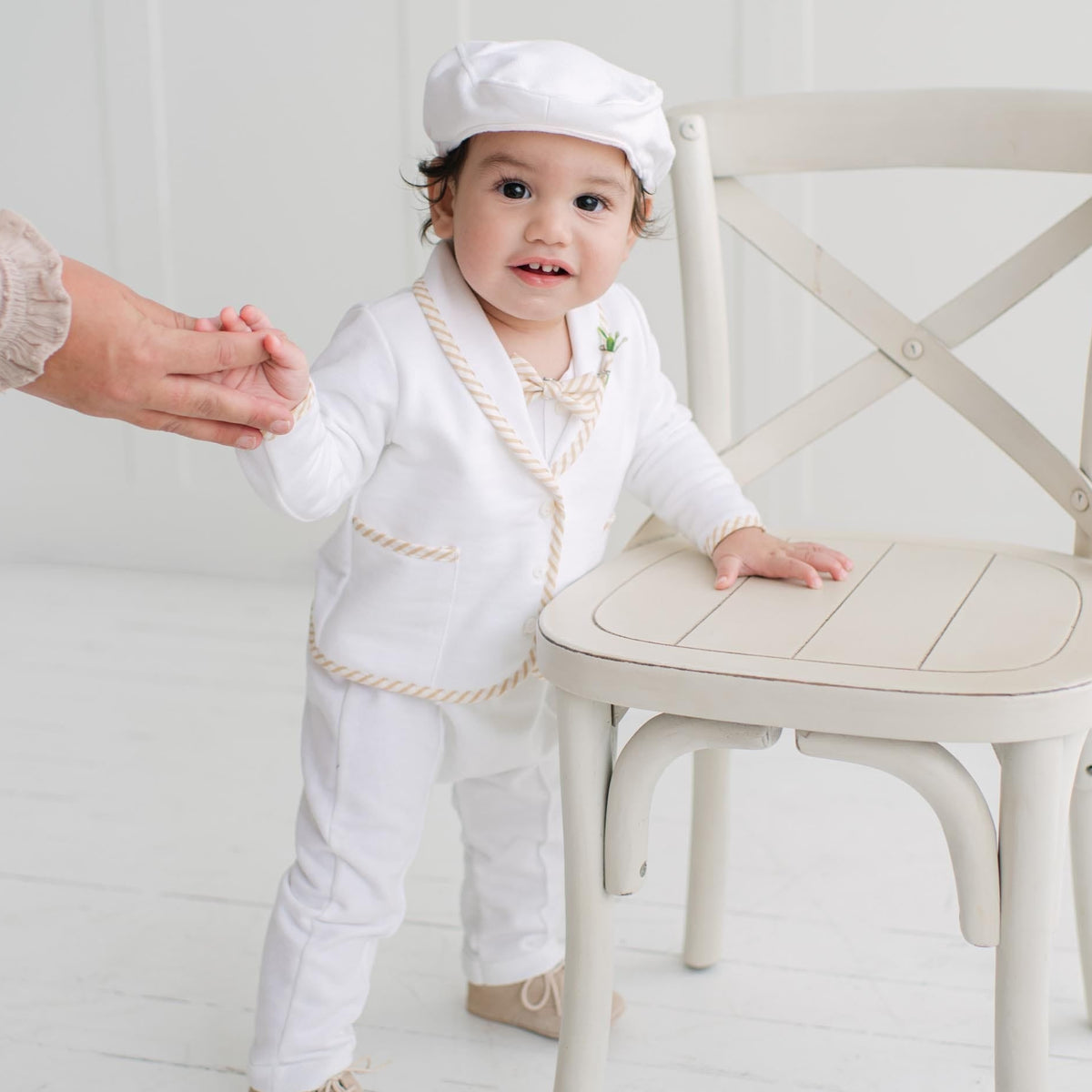 A toddler wearing a white baby outfit with beige striped trim and a matching white newsboy hat stands beside a cream-colored wooden chair, holding an adult's hand in a bright, white room.
