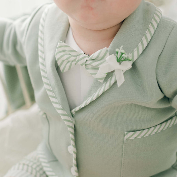 A close-up of a baby wearing the Theodore Bow Tie & Boutonniere suit. The light green suit, handmade in the USA, features white and green striped cotton trim and a matching bow tie. White buttons adorn the front, and there is a small floral boutonnière on the left lapel. The baby's face is partially visible.