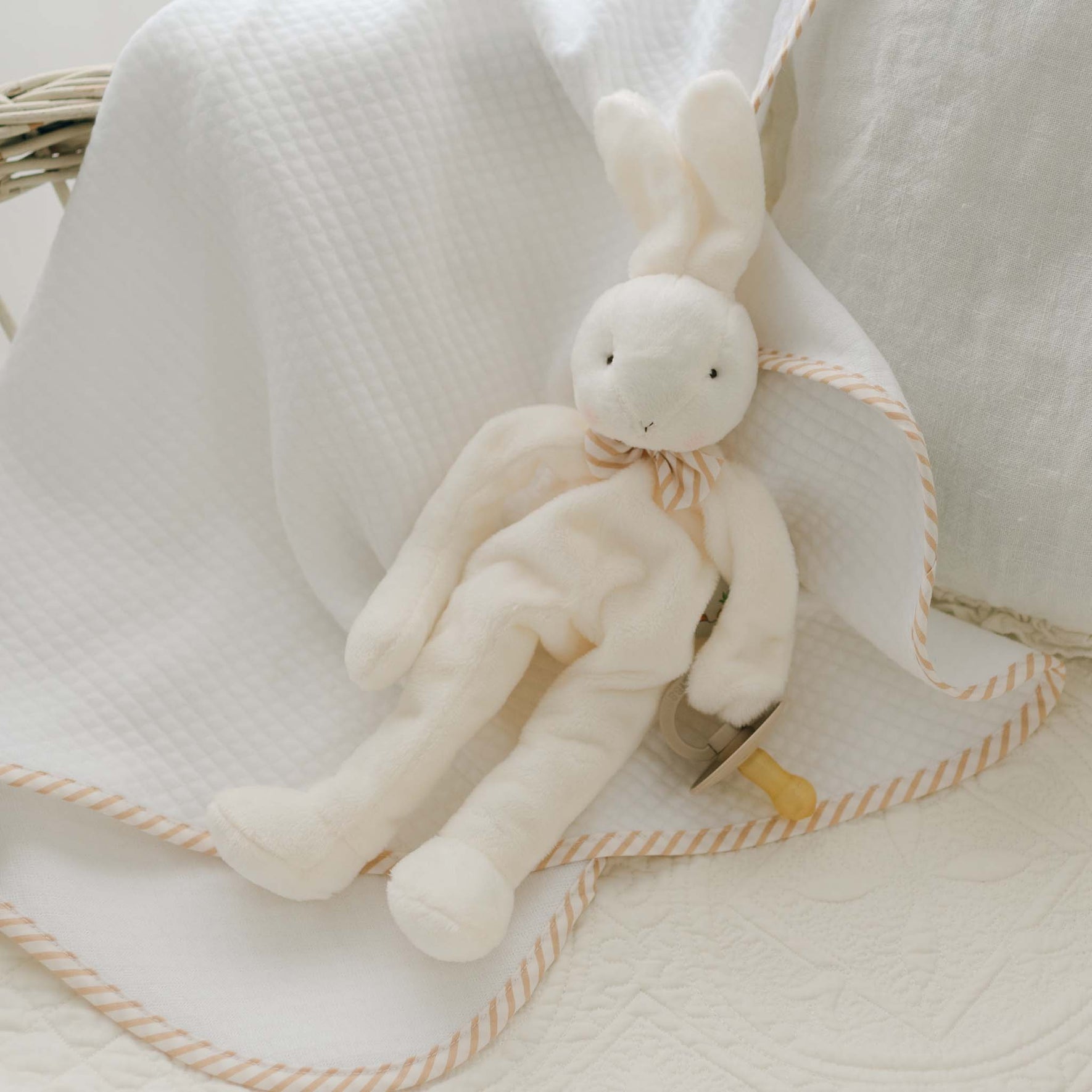 A Theodore Bunny Buddy | Pacifier Holder, featuring a white stuffed bunny with a striped bow around its neck, rests on a white quilted blanket with a striped border. The pacifier clip is tucked beneath the bunny's arm, arranged on the blanket. This charming setup seems to be in a cozy, baby-safe nursery corner or lovingly prepared crib—an ideal christening gift.