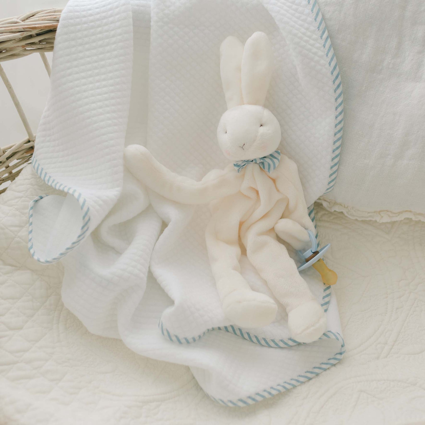 The Theodore Bunny Buddy Pacifier Holder, adorned with a blue and white striped bow around its neck, rests on a white blanket featuring blue striped trim. Perfect as a christening gift, the blanket and toy are placed on a cushioned surface, creating a cozy and soft appearance.