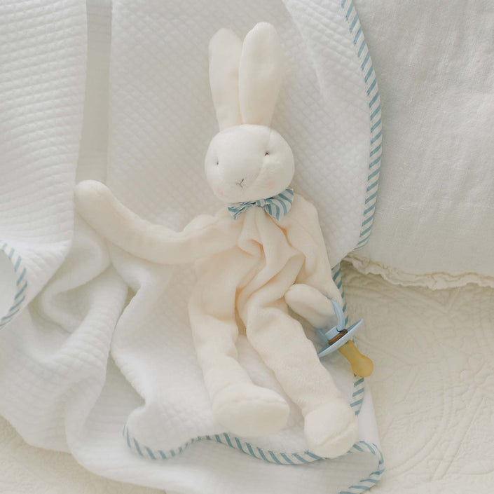 The Theodore Bunny Buddy Pacifier Holder, adorned with a blue and white striped bow around its neck, rests on a white blanket featuring blue striped trim. Perfect as a christening gift, the blanket and toy are placed on a cushioned surface, creating a cozy and soft appearance.