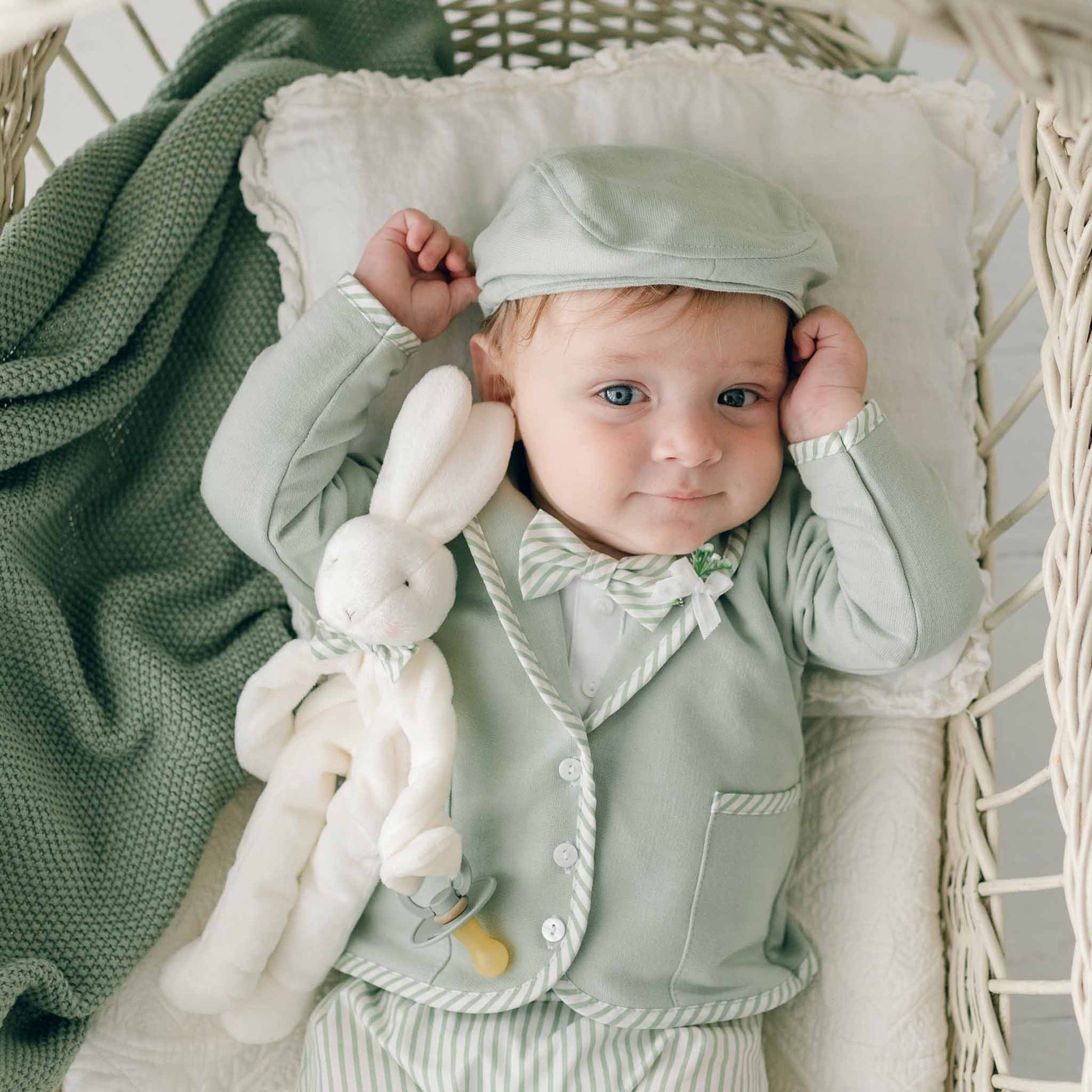 A baby dressed in light green clothing, including a hat and bow tie, lies on a white cushion in a wicker basket. The baby is holding the Theodore Bunny Buddy | Pacifier Holder with one hand and has a green blanket partially covering the basket – an adorable scene perfect as inspiration for a Christening gift.