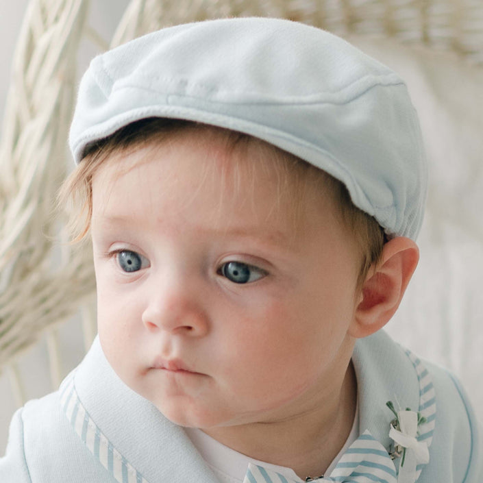 A baby with blue eyes is wearing the Theodore Newsboy Cap in light blue, accompanied by a matching outfit made of soft French terry cotton that features a white collar with blue stripes. The baby gazes to the left with a neutral expression against a blurred backdrop of wicker furniture or similar decor.