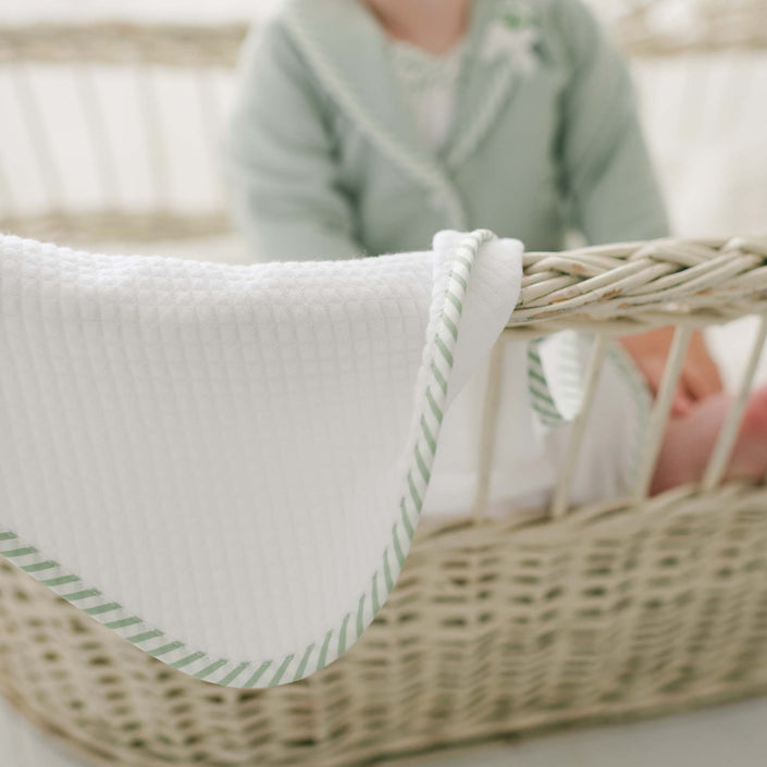 A baby dressed in a light green outfit is seated in a wicker bassinet. The focus is on the Theodore Personalized Blanket—a white blanket with green striped trim—draped over the edge of the bassinet. The background is softly blurred.