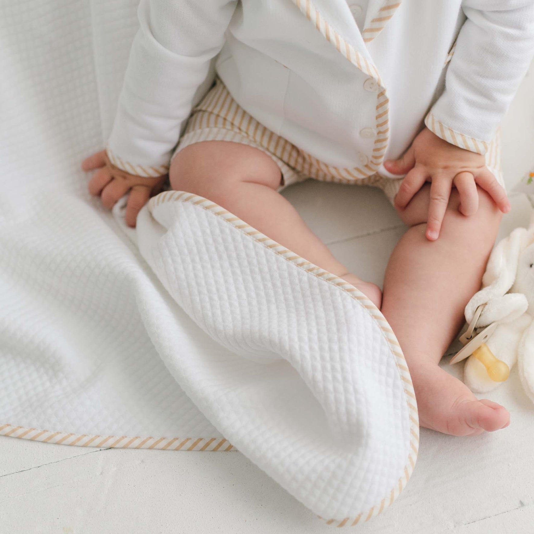 A baby is sitting on the Theodore Personalized Blanket, which features tan stripes. The baby is wearing a white outfit with matching tan stripe details. The baby's left leg is relaxed while the right hand rests on the thigh. A soft toy is partially visible next to the baby, creating a perfect scene for a personalized baby gift or christening gift photo.