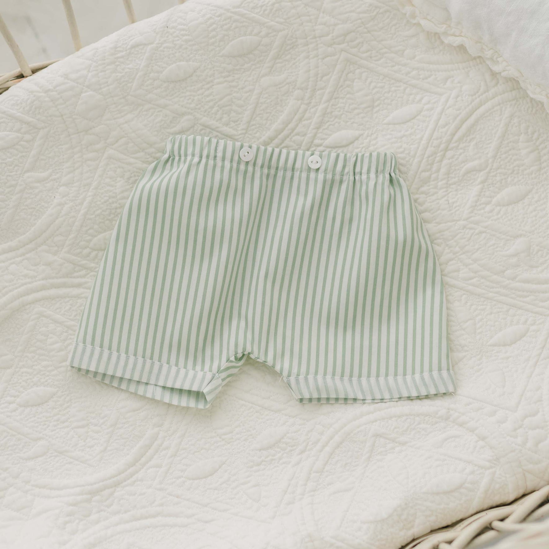 The Theodore Shorts Suit, a handmade baby boy outfit featuring light green and white striped shorts with an elastic waistband and white buttons, is displayed on a quilted, white patterned blanket. The suit includes white trim at the leg openings and is ideal for special occasions.