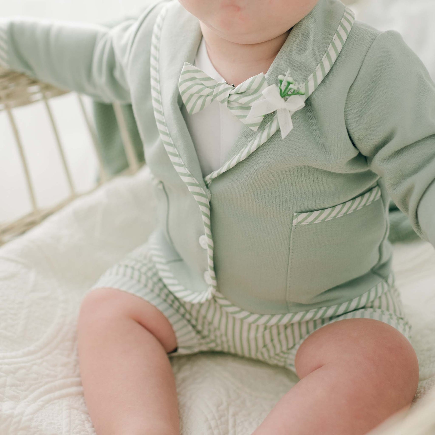 A baby wearing the Theodore Shorts Suit, a pastel green outfit featuring a jacket with white trim, a matching bow tie, and striped shorts, sits on a light-colored quilt. The baby's face is only partially visible, emphasizing the clothing's details. This handmade suit is perfect for special occasions!