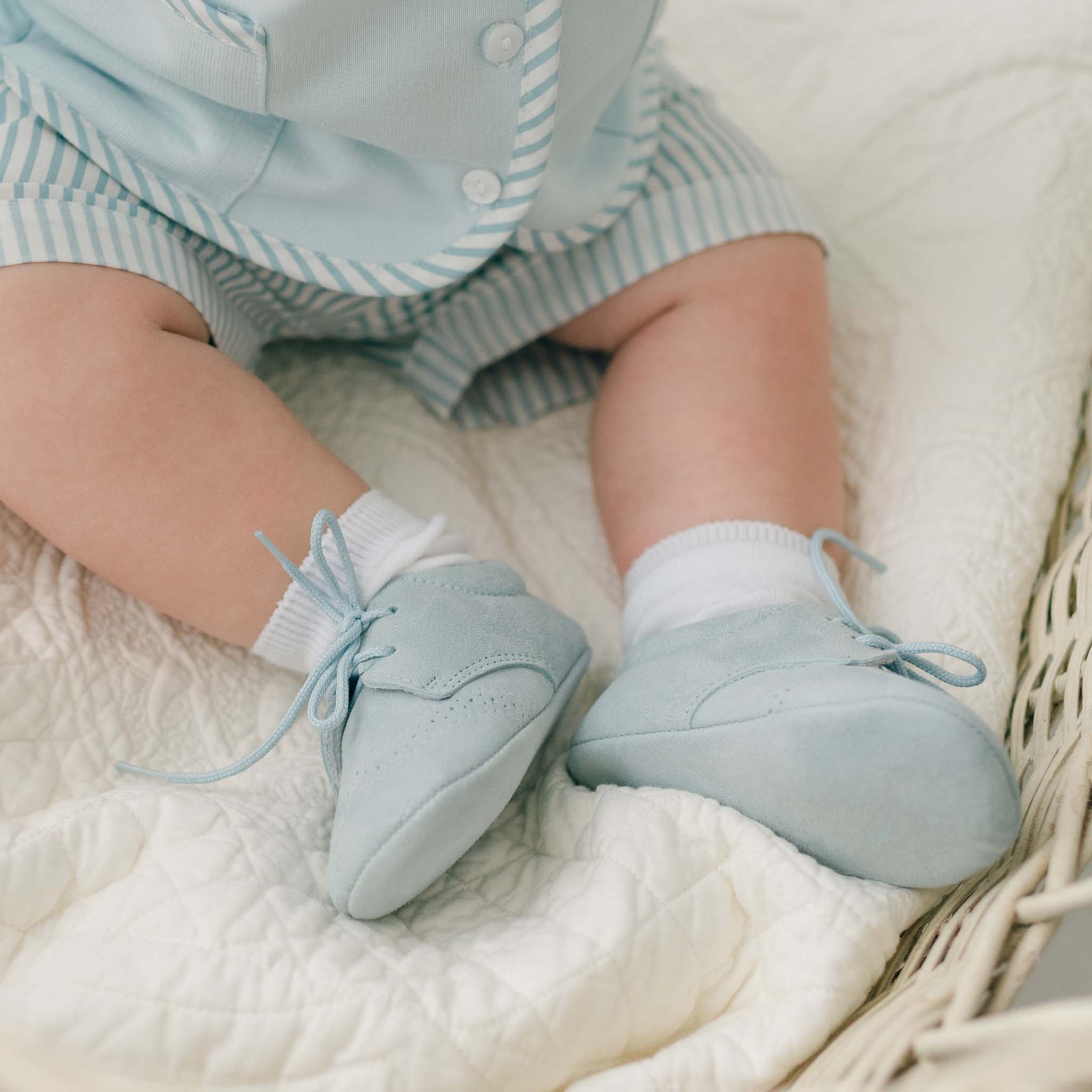 A baby is sitting on a quilted blanket, wearing a light blue outfit with striped shorts. The baby's feet are dressed in dove grey suede Theodore Suede Shoes by Baby Beau & Belle, paired with white socks. Handmade in Spain, the child is positioned in a wicker basket or bassinet.