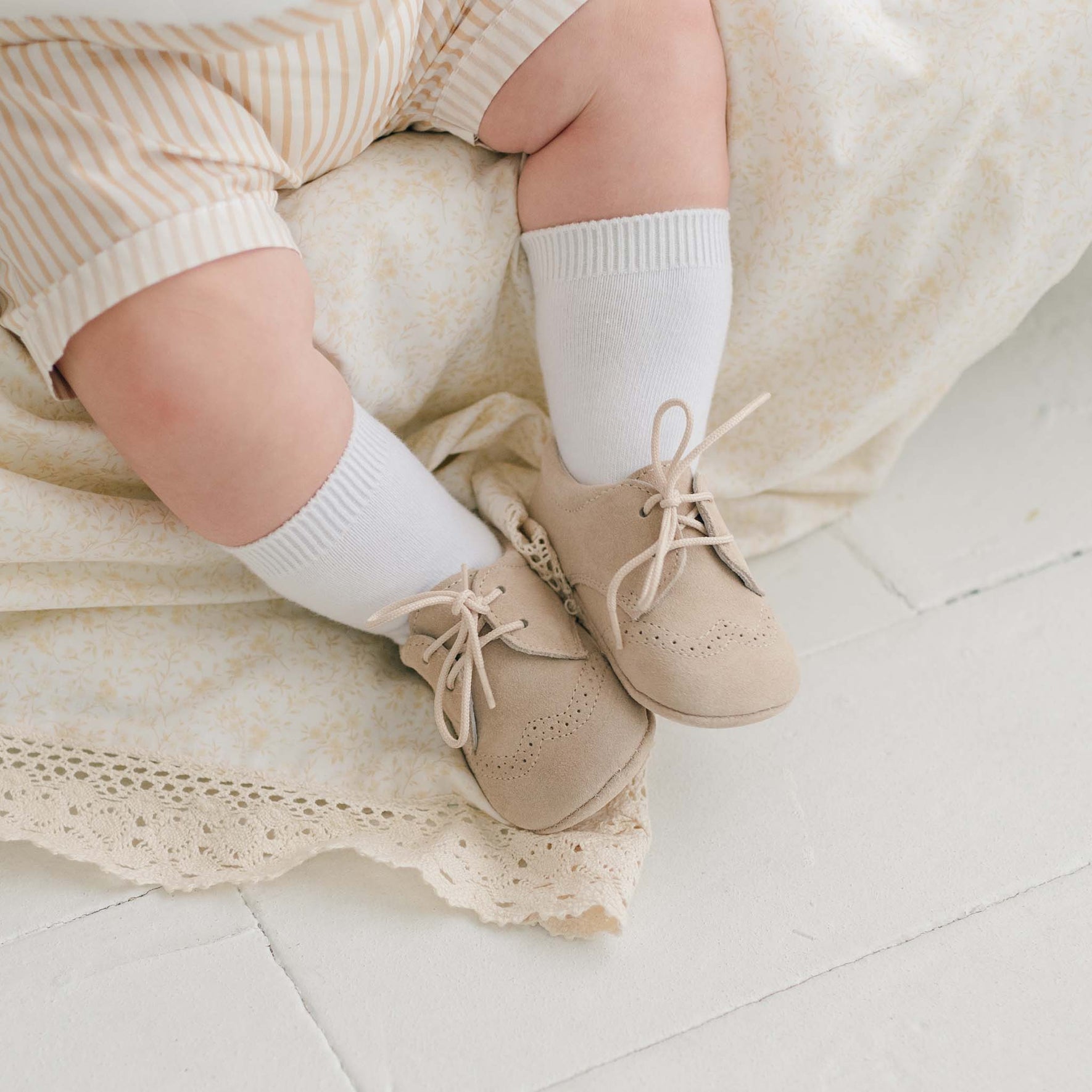 A baby's legs and feet are shown wearing Theodore Suede Shoes in dove grey. The baby is dressed in light-colored, striped clothing and sits on a cream-colored lace-trimmed blanket. The background is a white tiled floor.