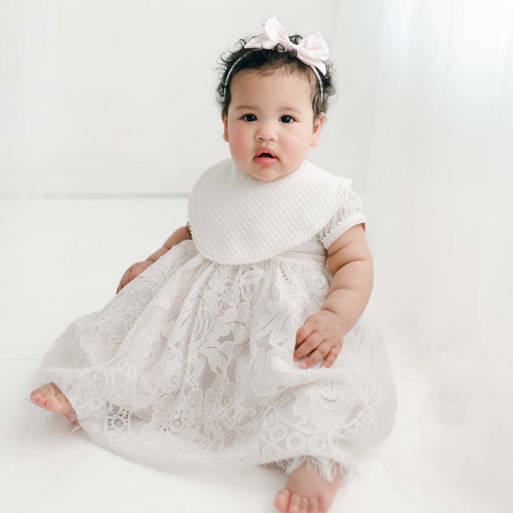 A baby with curly hair sits on the floor wearing the Victoria Christening Bib, over the Victoria Puff Sleeve Christening Dress, and the Victoria Silk Bow Headband.